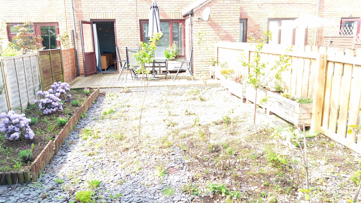 The rear garden features a wooden deck with outdoor seating, surrounded by blooming flowers and greenery. The area displays a mix of gravel and soil, providing a simple yet inviting space for relaxation, with access leading into the house through glass doors.