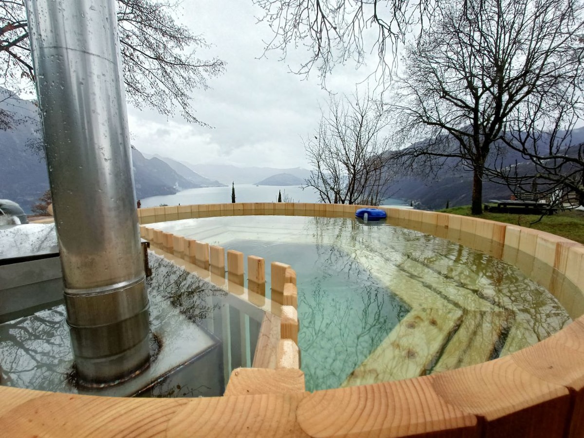 A wooden hot tub sits on an elevated deck overlooking a serene lake. The natural landscape is framed by trees and mountains in the distance, creating a tranquil environment. Steam rises gently from the water's surface, suggesting warmth and relaxation.
