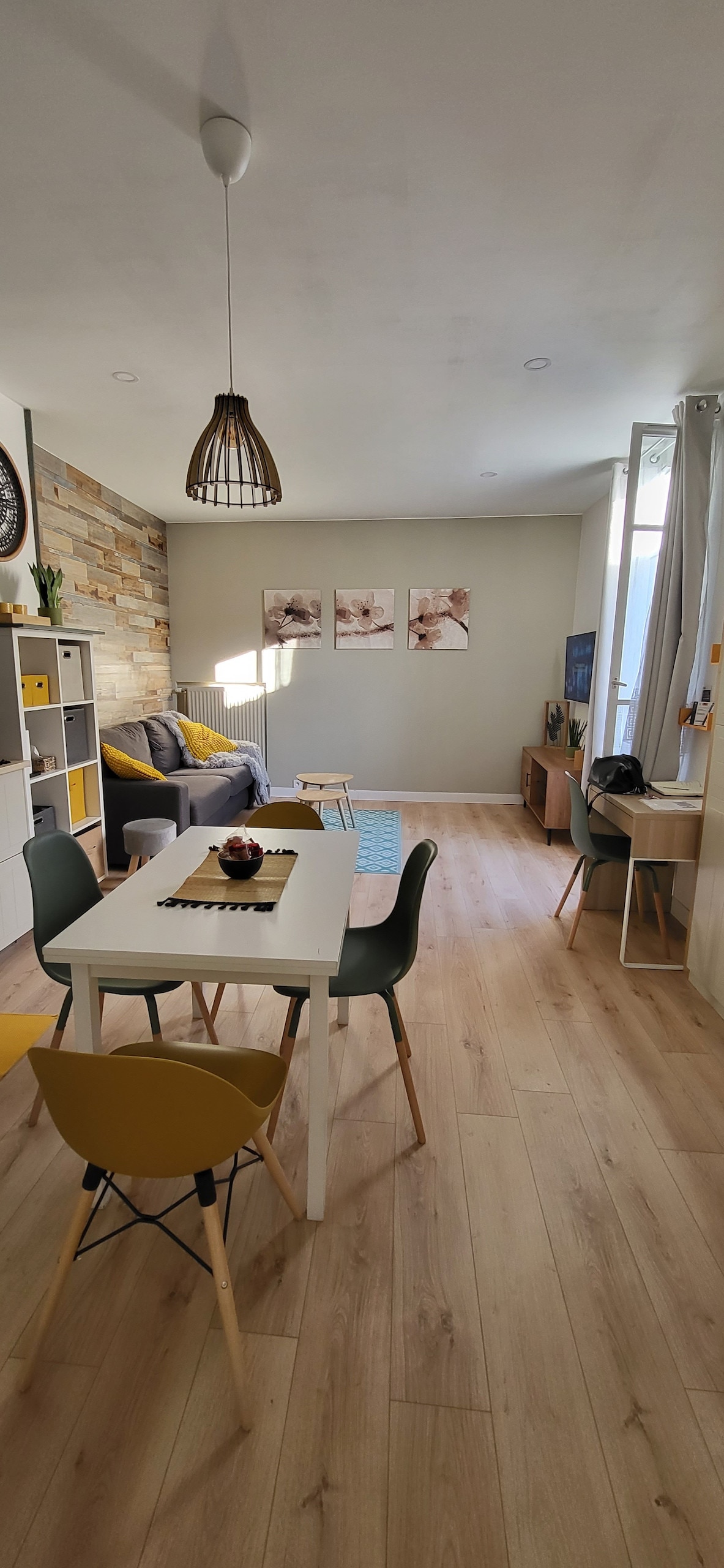 A modern and well-lit living area features a stylish table set for four, complemented by colorful chairs. A comfortable sofa faces a media console, with wall art adding subtle decor. Natural light pours in through large windows, enhancing the inviting atmosphere of the space.