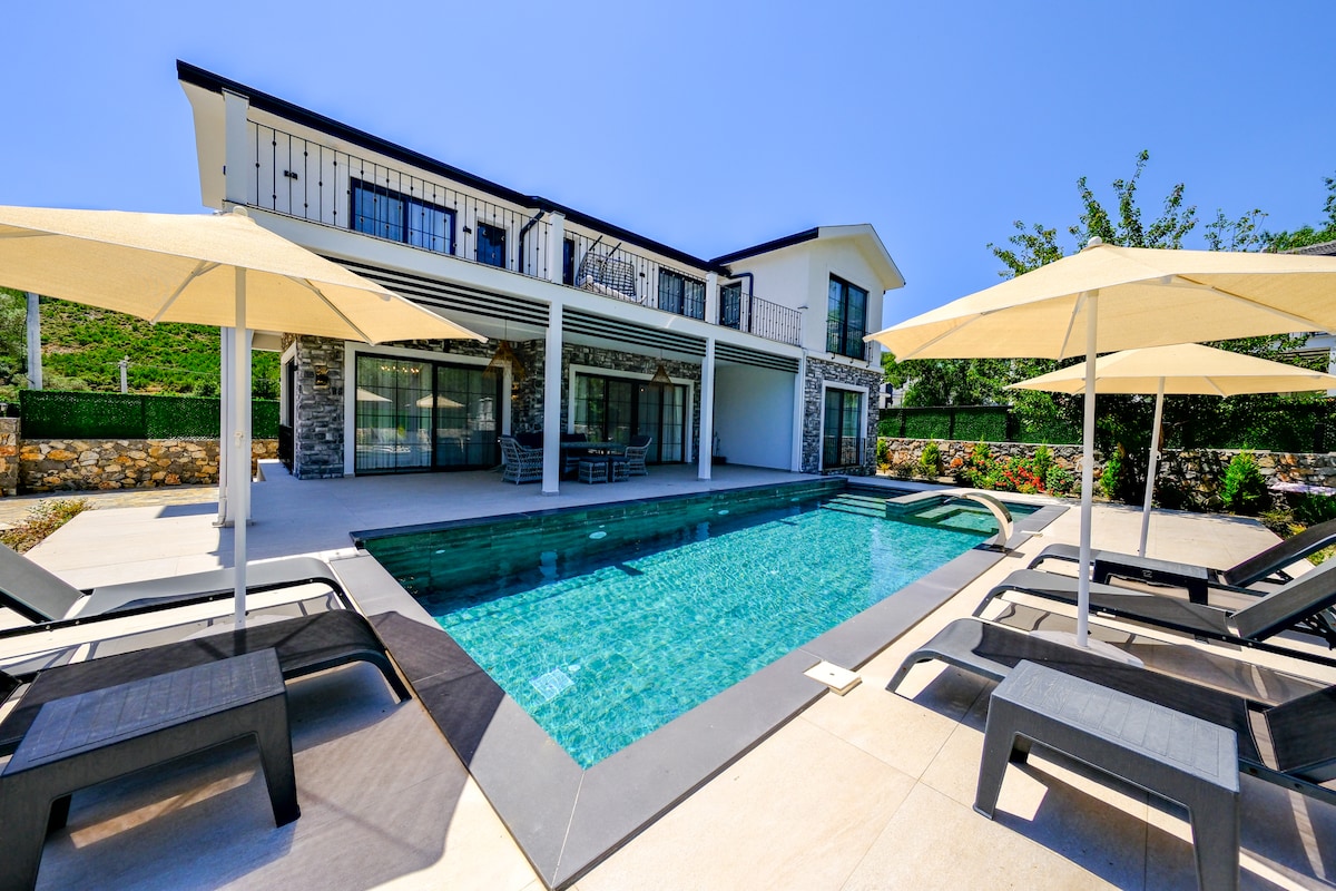 The exterior of the villa showcases a modern design with large glass doors leading to the pool area. A private swimming pool is surrounded by sun loungers and umbrellas, creating a refreshing and inviting outdoor space. The lush greenery enhances the serene atmosphere.