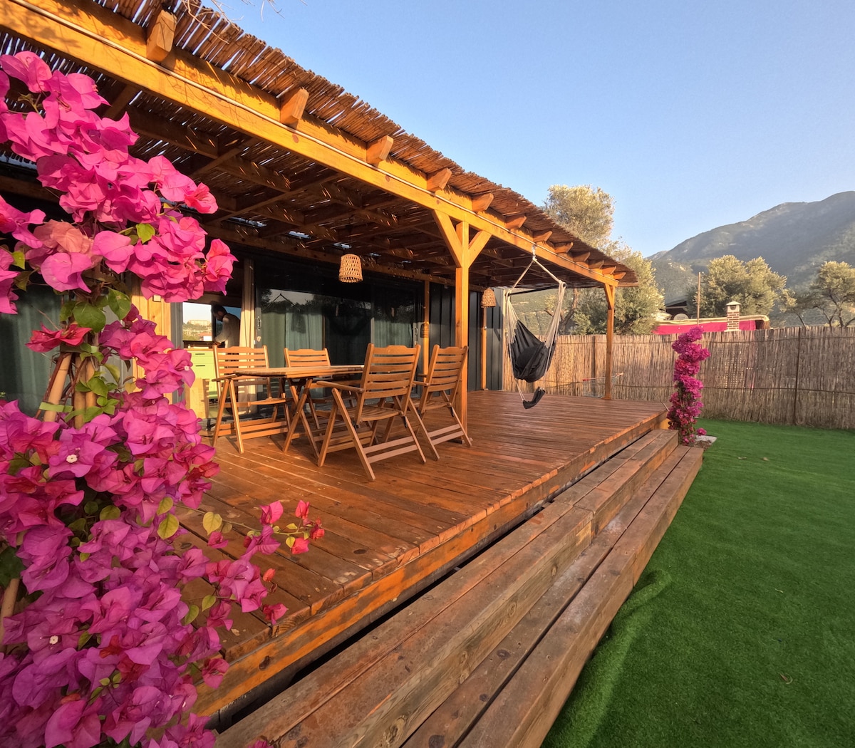 A spacious wooden terrace is adorned with vibrant pink bougainvillea flowers. The area features a dining set with wooden chairs and a hammock for relaxation. Natural beauty is complemented by views of surrounding greenery and mountains in the distance, providing a tranquil setting.