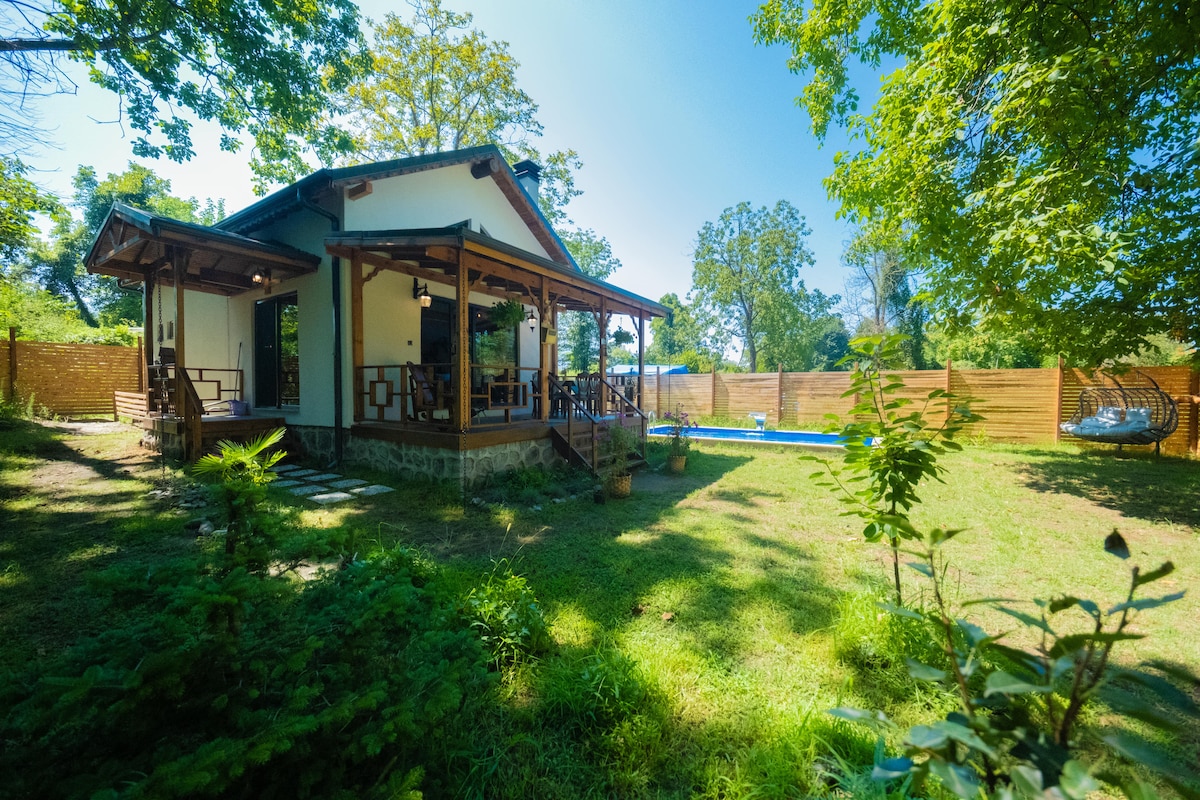 A charming bungalow is set in a lush green garden, surrounded by trees. The outdoor space includes a swimming pool and a covered porch with seating. The neutral-toned exterior complements the natural surroundings, while the private fencing provides a sense of seclusion.
