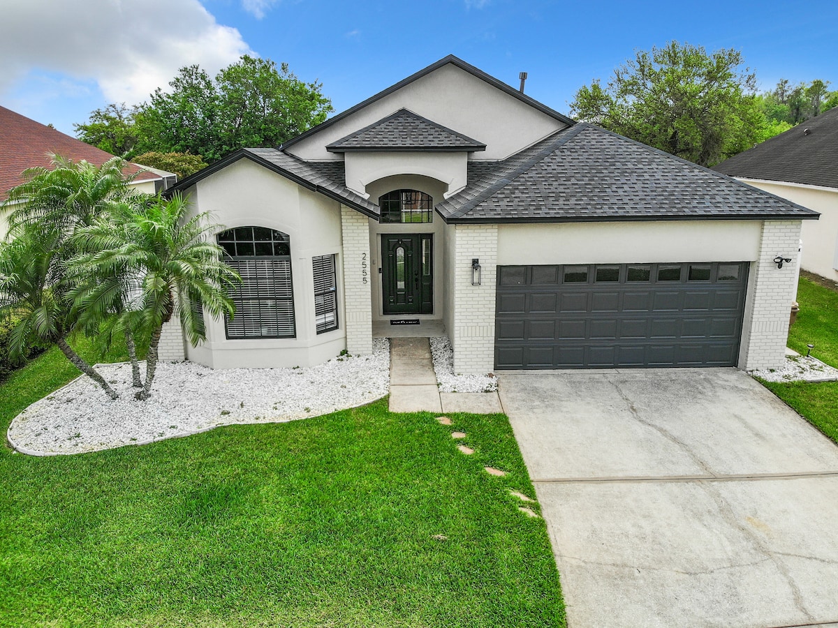 A modern, one-story home features a well-maintained lawn and palm trees. The exterior showcases a mix of light and dark elements, with a prominent driveway leading to a two-car garage. Large windows allow for ample natural light, contributing to the inviting atmosphere.