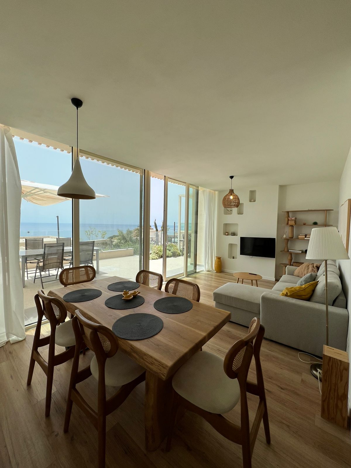 A dining area features a wooden table surrounded by eight chairs. A cozy seating arrangement is positioned nearby, facing a wall-mounted TV. Large glass doors provide a view of the outdoor space with ocean views, while natural light brightens the room through sheer curtains.