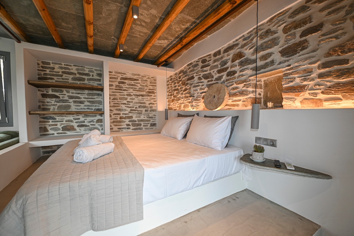 A cozy sleeping area is featured with a spacious bed adorned with soft white linens. Exposed stone walls add texture to the room, while integrated shelves provide practical storage. Natural light flows in from a nearby window, highlighting the modern, minimalist design.