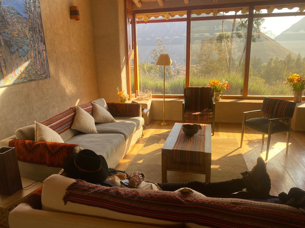 A sunny living area features large windows that frame views of the surrounding mountains. A soft couch and colorful armchairs are arranged around a wooden coffee table. Natural light enhances the textured walls, while floral arrangements add a touch of warmth to the space.
