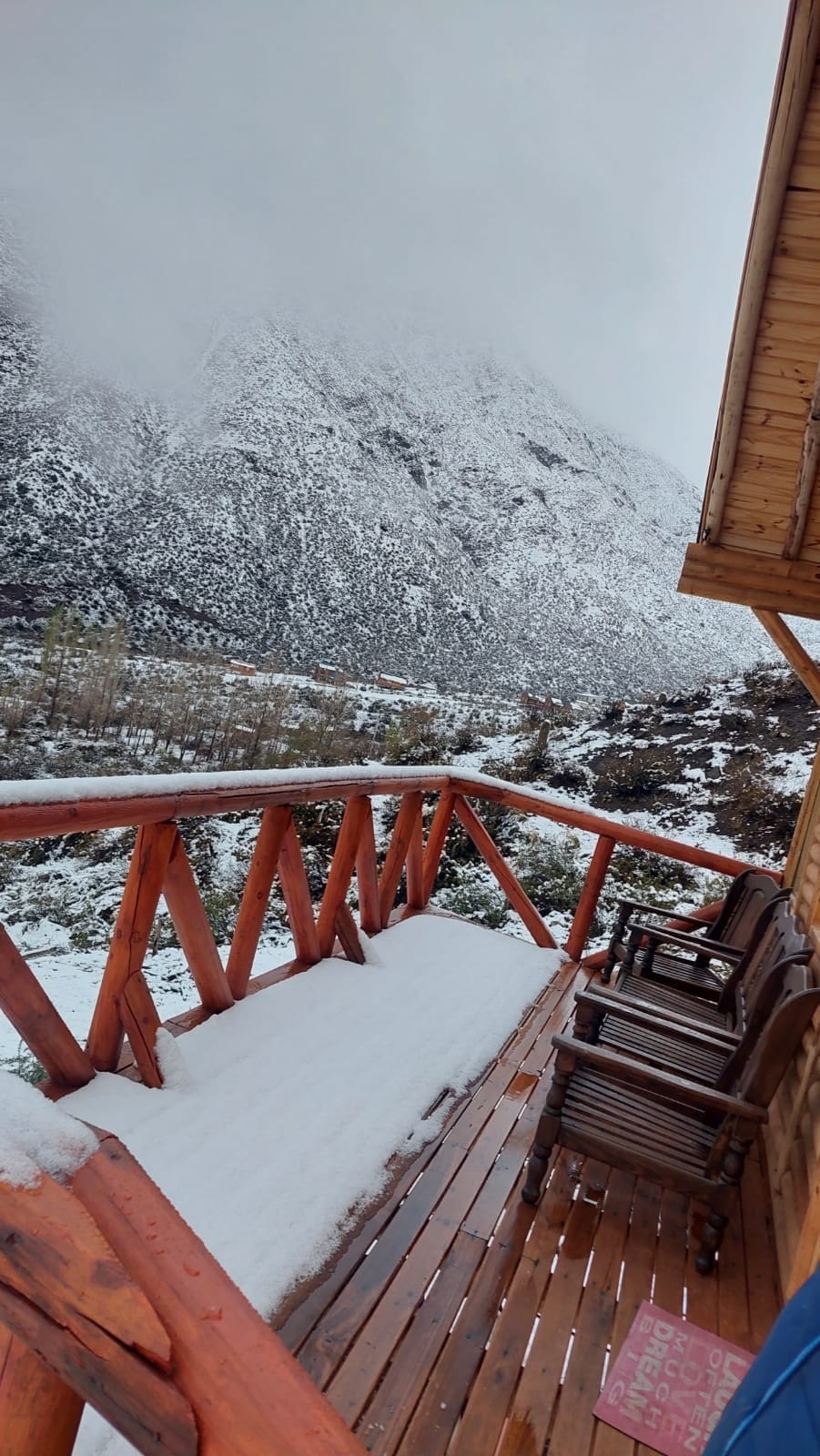 A wooden deck extends from the cabin, covered in a light layer of snow. Several outdoor chairs are arranged along the railing, offering a view of snow-capped mountains shrouded in mist and clouds.