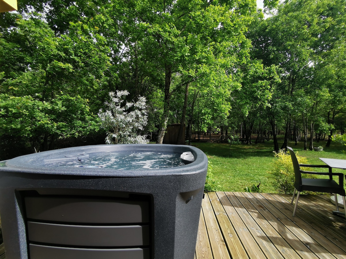 A private spa is situated on a wooden terrace, surrounded by lush greenery. The view includes trees and a well-maintained garden, creating a serene outdoor environment. A dining area with chairs is observable in the foreground, contributing to the relaxing ambiance.