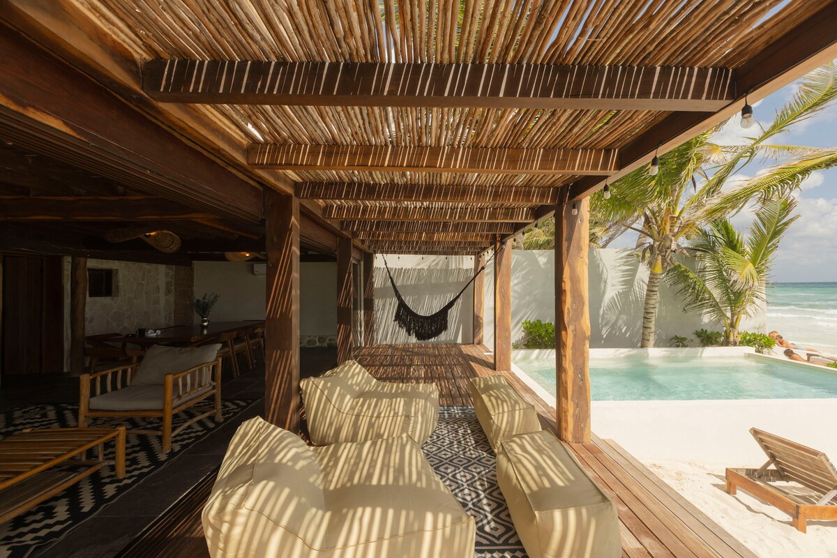 An outdoor terrace features a wooden structure with a natural thatched roof, providing shade over several lounge seating options. A cozy hammock is suspended nearby, while a clear blue pool is visible in the background, along with palm trees swaying gently by the beach.