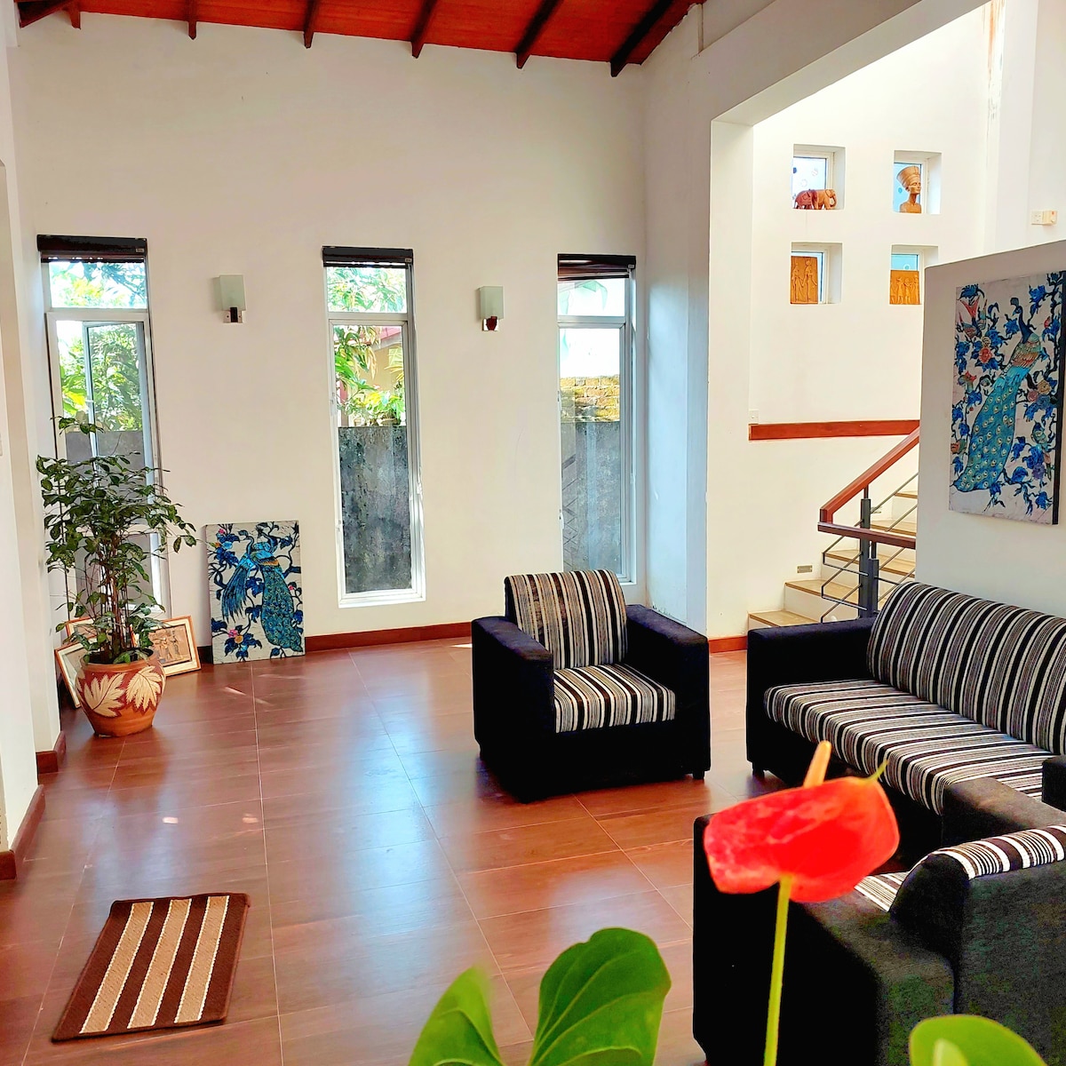 A bright living area features large windows providing natural light. Two striped armchairs and a spacious striped sofa are arranged around a central space. A potted plant adds a touch of greenery, while colorful wall art complements the neutral walls and tiled flooring.