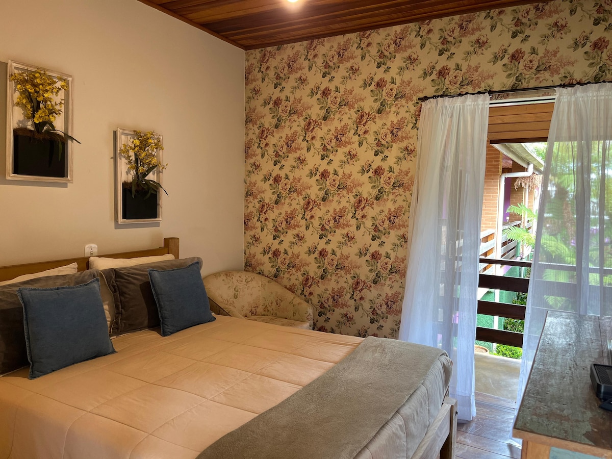 A cozy sleeping area features a spacious bed with soft linens, complemented by decorative pillows. Floral wallpaper adds charm, while natural light enters through a sheer curtain. A small seating area is visible, enhancing the room's inviting atmosphere.