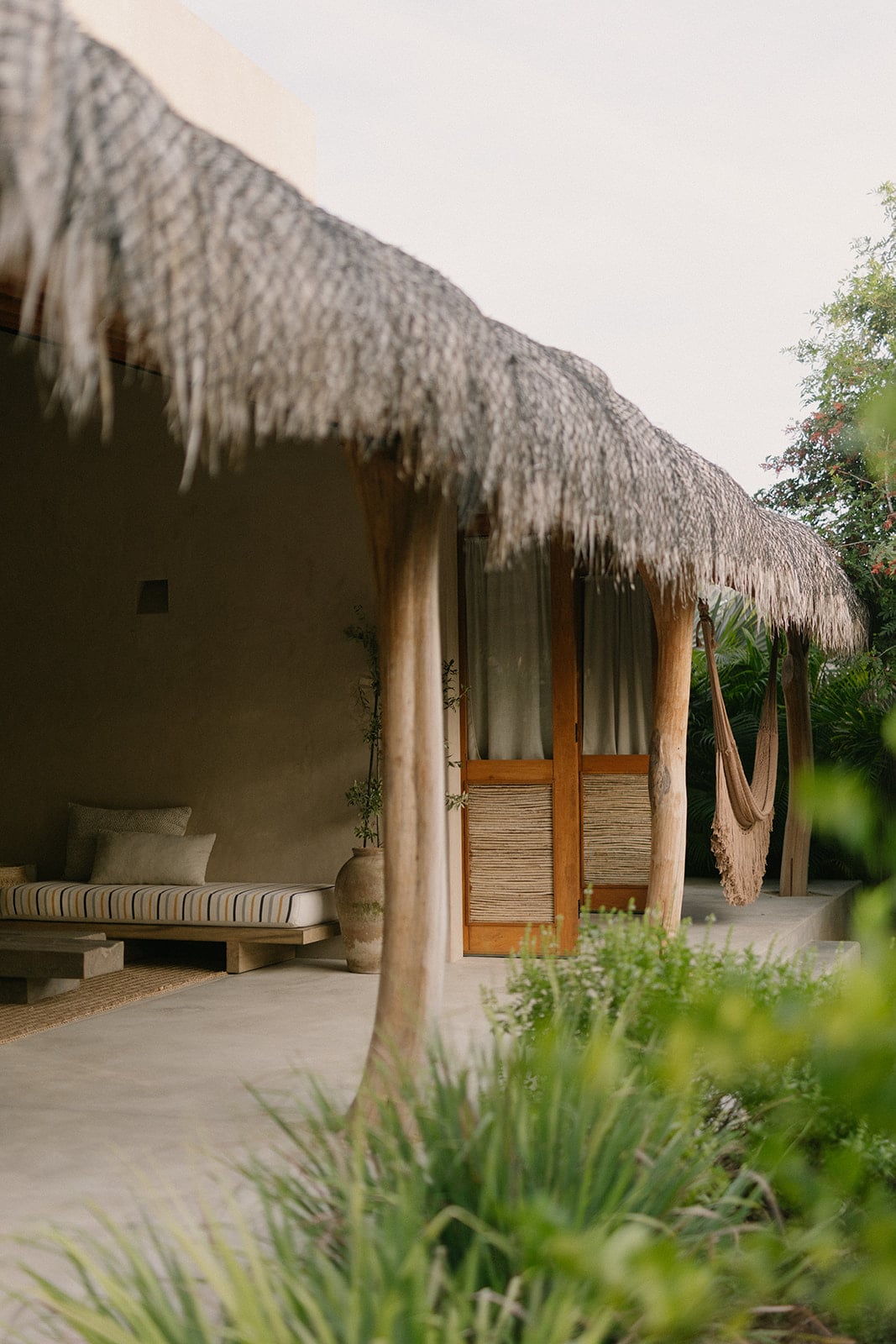 A rustic structure with a thatched roof is observed, featuring wooden doors with a natural finish. A comfortable seating area with striped cushions is positioned nearby, accompanied by a hammock. Lush greenery and plants create a serene environment, enhancing the outdoor space.