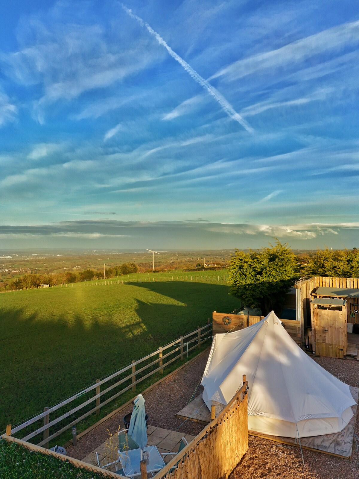 A 5m bell tent is situated in a private area, with lush green fields stretching into the distance. The sky is filled with wispy clouds and a light blue hue, while shadows from nearby trees and structures create a serene landscape.