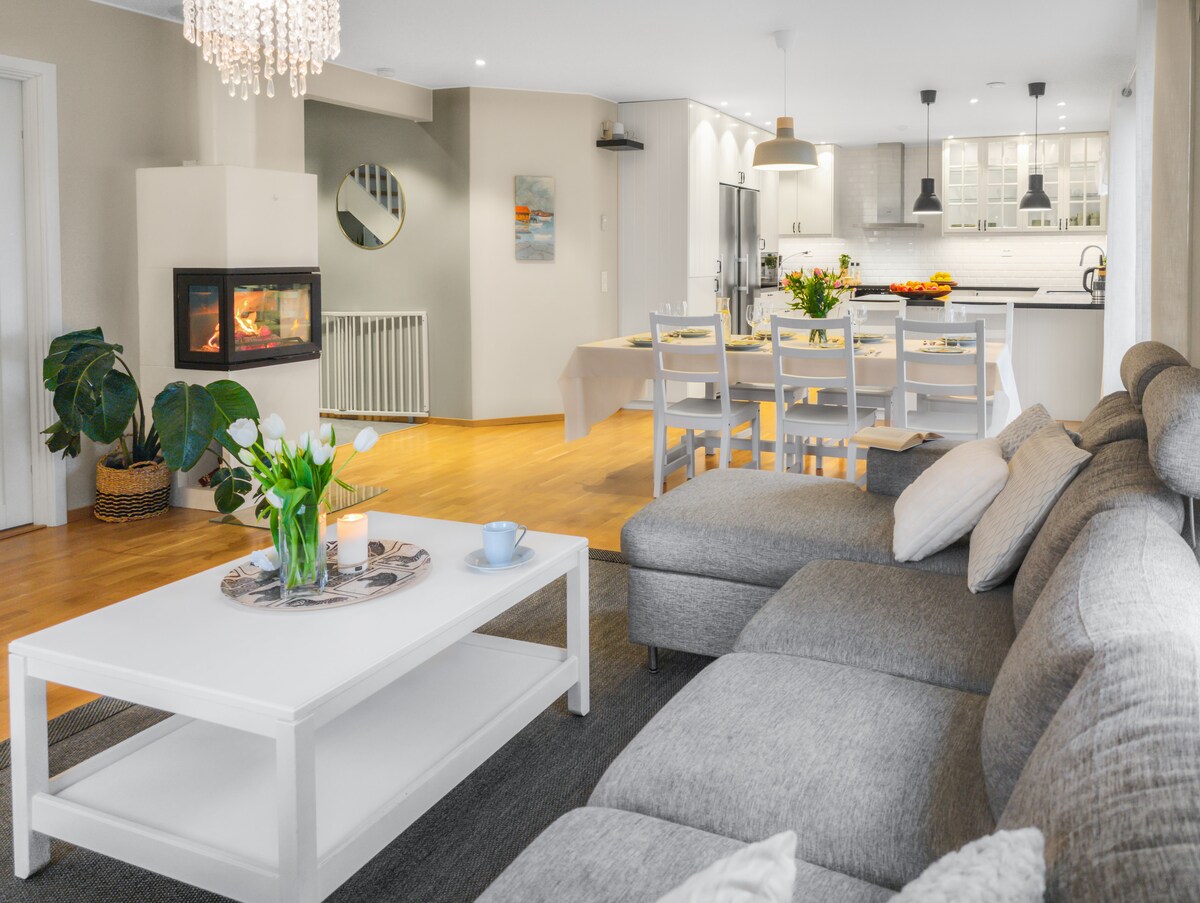 A spacious living area features a comfortable gray sectional sofa arranged around a white coffee table. In the background, a modern kitchen is visible, complemented by a dining table set for eight and bright countertops. A fireplace adds warmth to the inviting atmosphere.