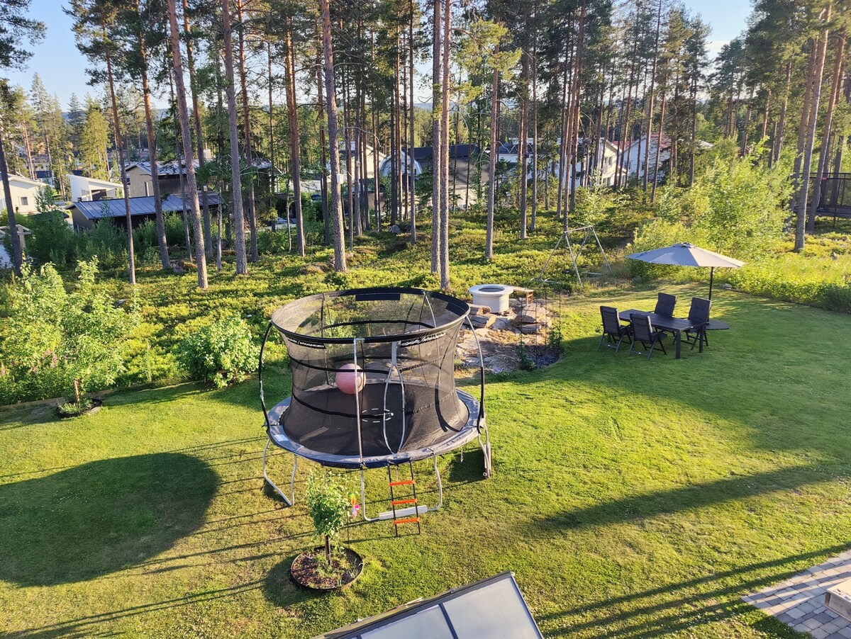 A spacious garden is seen, featuring a large trampoline surrounded by green grass and trees. A fire pit area with seating is visible, along with a shaded dining space under a canopy. The natural surroundings enhance the outdoor experience.