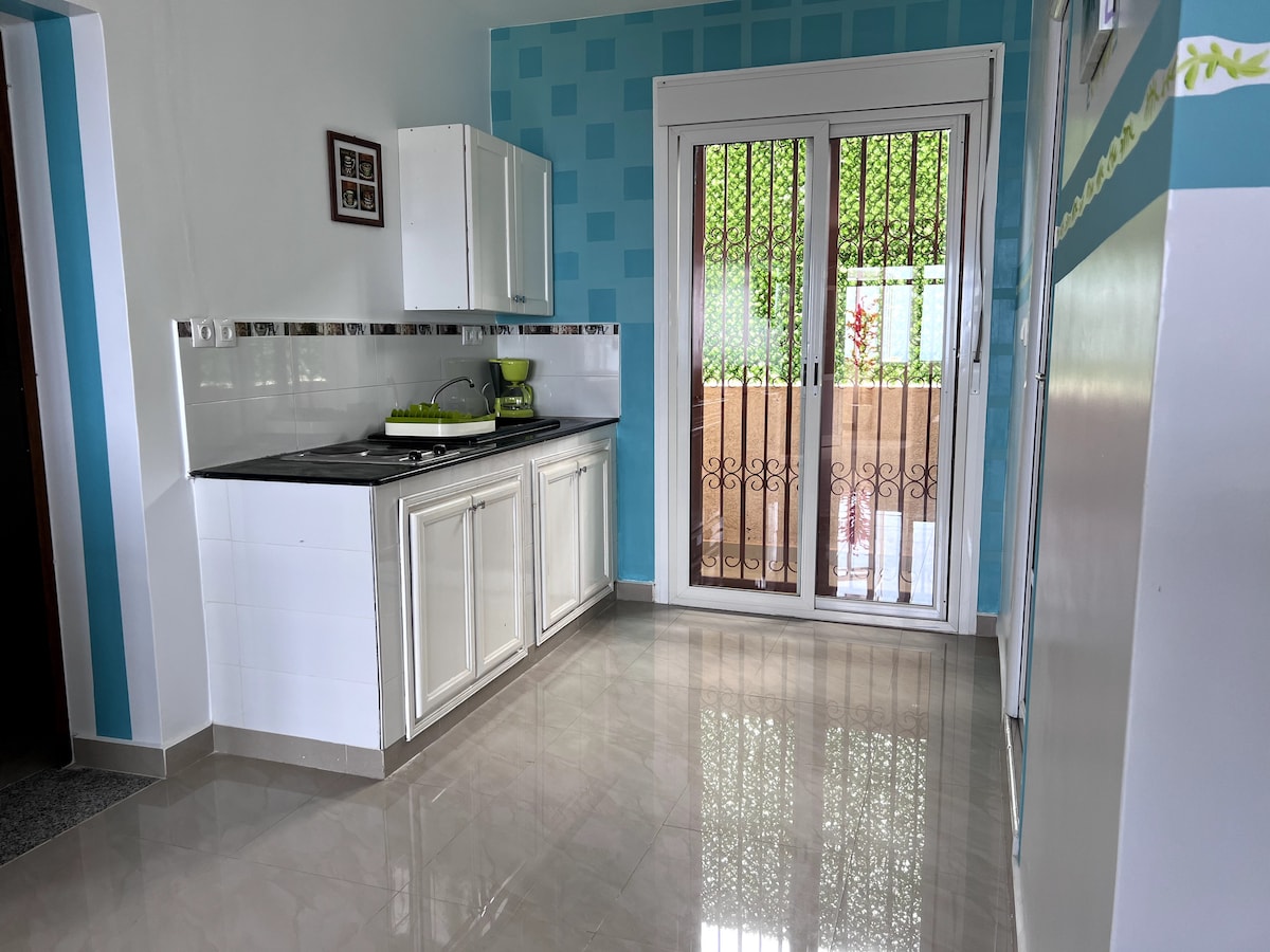 A bright kitchen area features white cabinetry and a black countertop. Light-colored tiles reflect natural light, enhancing the space. A glass door with decorative bars opens to a private outdoor area, surrounded by greenery. Colorful accent walls complete the cheerful atmosphere.