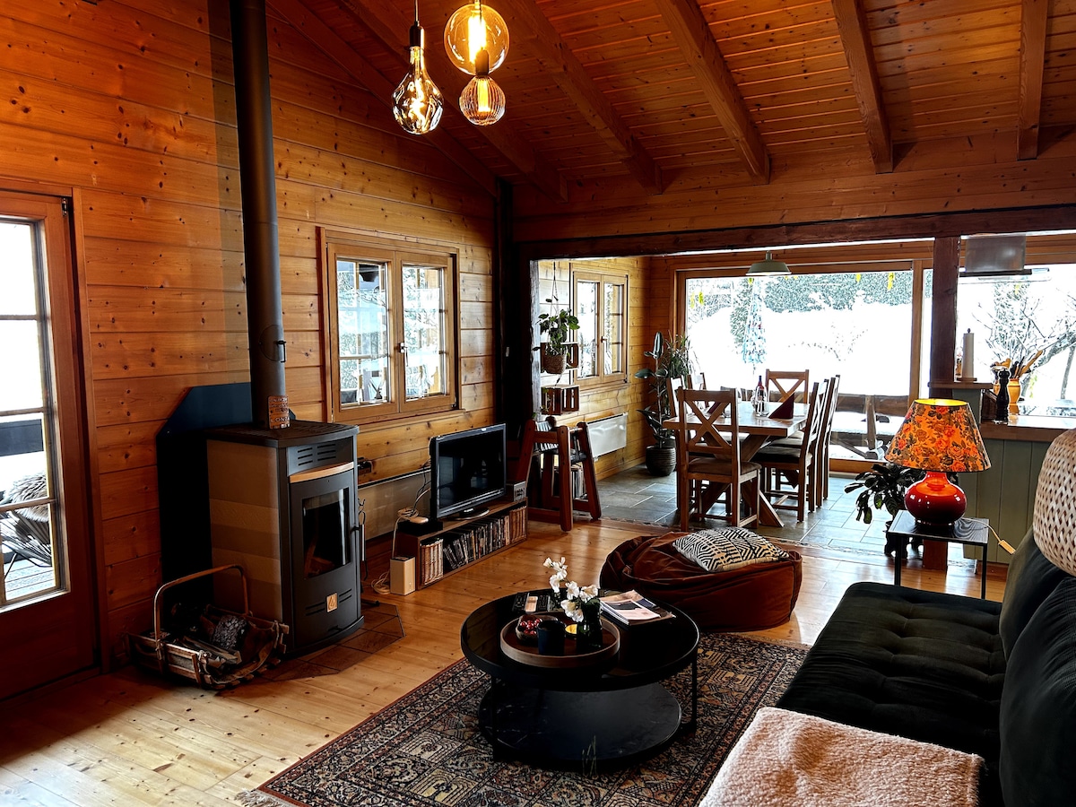 A wooden interior features high ceilings with exposed beams, and large windows allowing natural light. A cozy seating area includes a dark sofa and a round coffee table. A dining area with wooden chairs and a table is visible, alongside a stylish lamp casting warm light.
