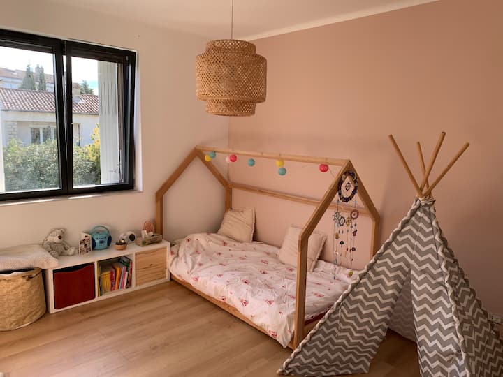 Veľká detská spálňa s hračkami, knihami a malým stolom. K dispozícii je úložná skriňa s vešiakmi, ventilátor a spálňa je vybavená okenicami a závesom. 
Je možné pridať nafukovací matrac s rozmermi 140