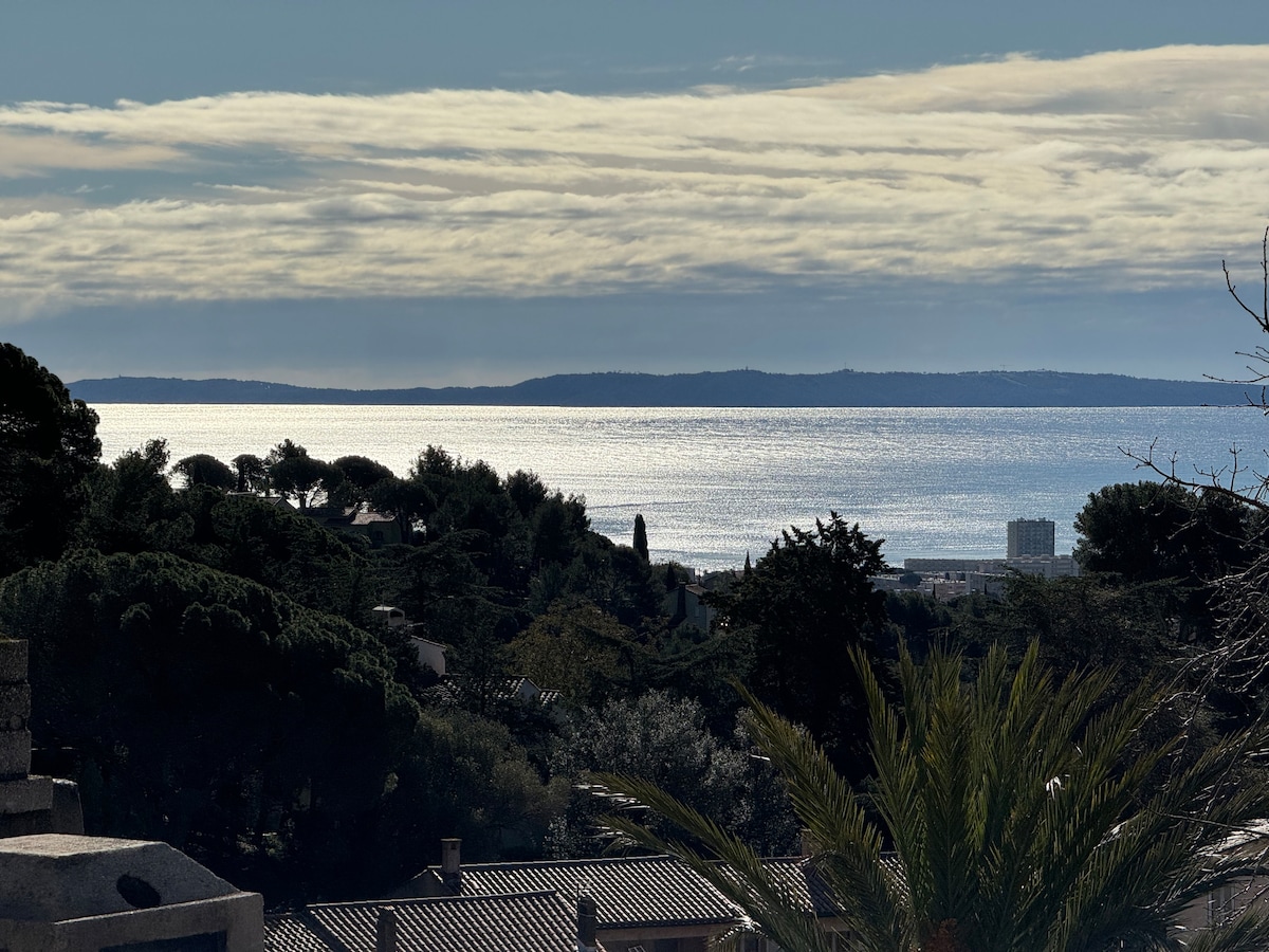 A serene view showcases the coastline reflecting sunlight on calm waters, framed by lush greenery. The horizon features distant islands under a gently clouded sky, conveying a sense of tranquility and connection with nature.