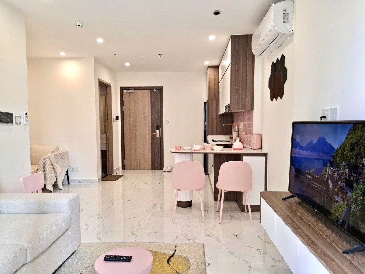 An open living space features modern furnishings, including a light-colored sofa and pink chairs. A sleek kitchen counter is visible in the background, with essential appliances and decorative items. Soft lighting enhances the contemporary design, while a flat-screen TV is placed on a wooden media console.