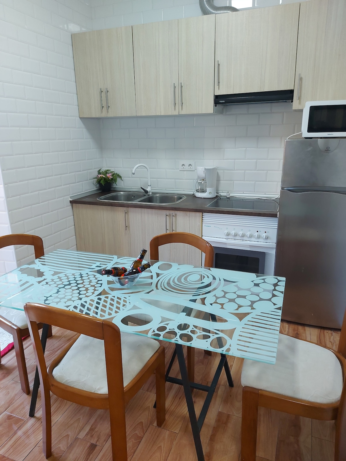 A modern kitchen area is showcased, featuring a glass dining table surrounded by four wooden chairs. Stainless steel appliances, including a refrigerator and oven, are present, alongside a sink and coffee maker. White tiled walls enhance the clean and bright atmosphere.