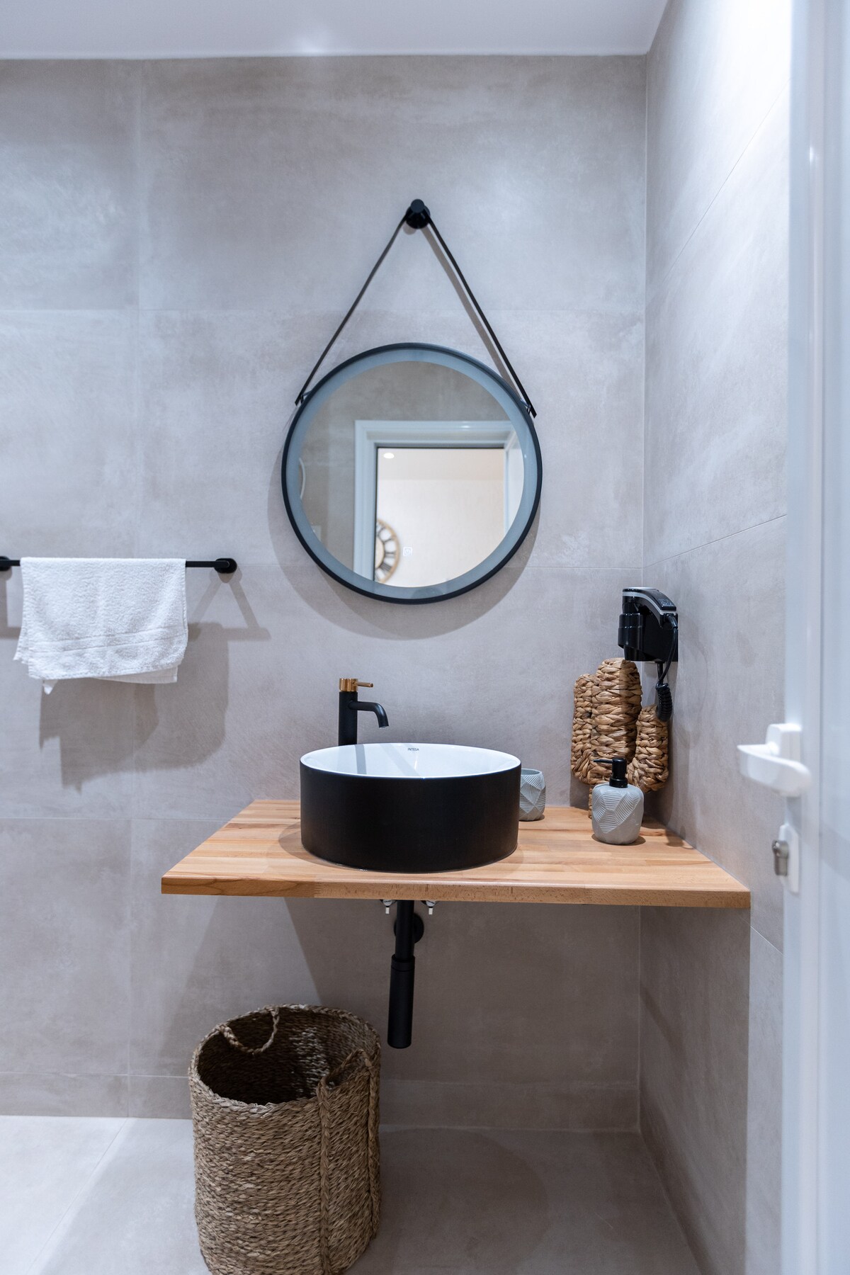 A modern bathroom space features a circular mirror above a sleek black basin set on a wooden countertop. Textured walls in a neutral tone complement a woven basket and decorative storage containers, providing an organized look. A towel bar and a fresh towel add practicality.