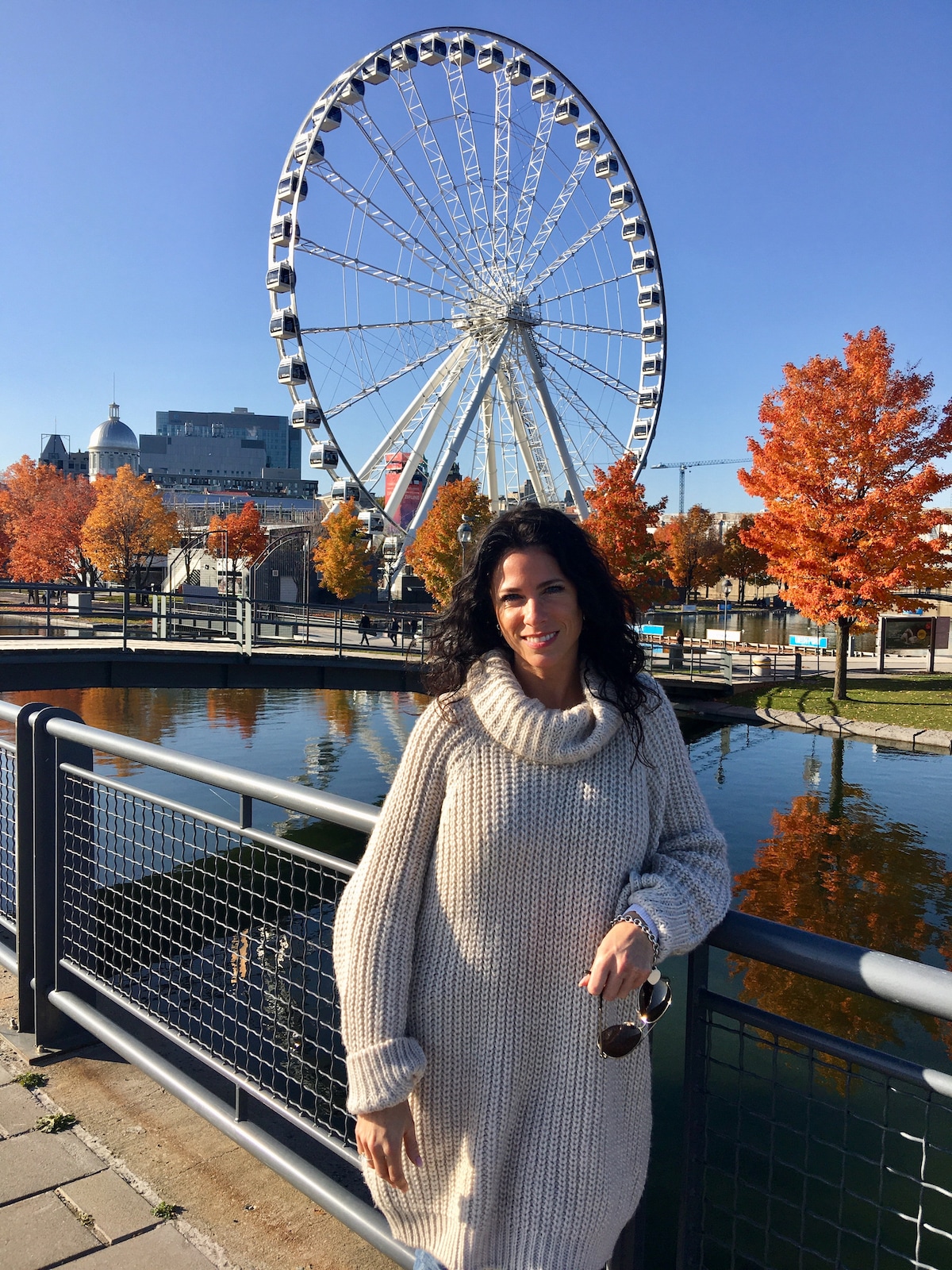 A large Ferris wheel stands prominently in the background against a clear blue sky. Autumn trees with vibrant orange leaves frame a peaceful waterway in the foreground, reflecting the surrounding colors and structures.