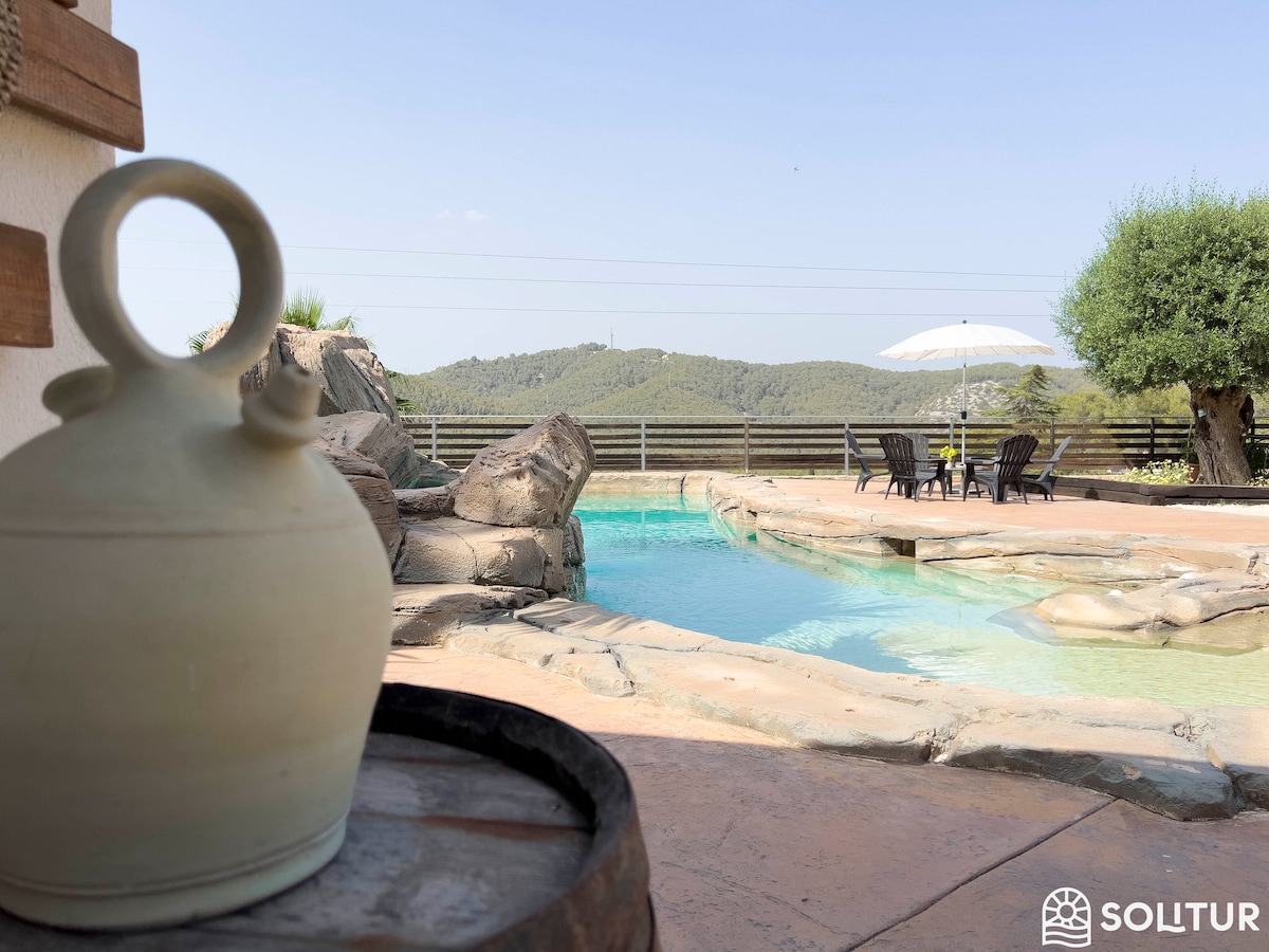 A serene pool area features a natural stone design, with a gentle curve mimicking a beach. A shaded umbrella provides respite from the sun, while a wooden barrel and ceramic jug accentuate the outdoor space. Lush green hills can be seen in the background.