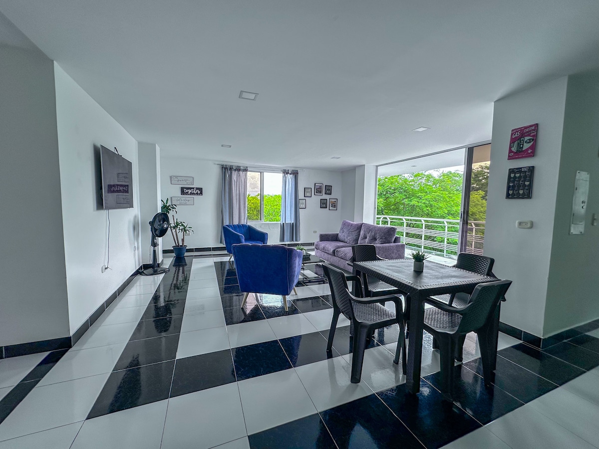 A bright living area features large windows allowing ample natural light to illuminate contrasting black and white tile flooring. Comfortable seating is arranged with purple sofas and a dining table surrounded by chairs, complemented by stylish wall decor and indoor plants.