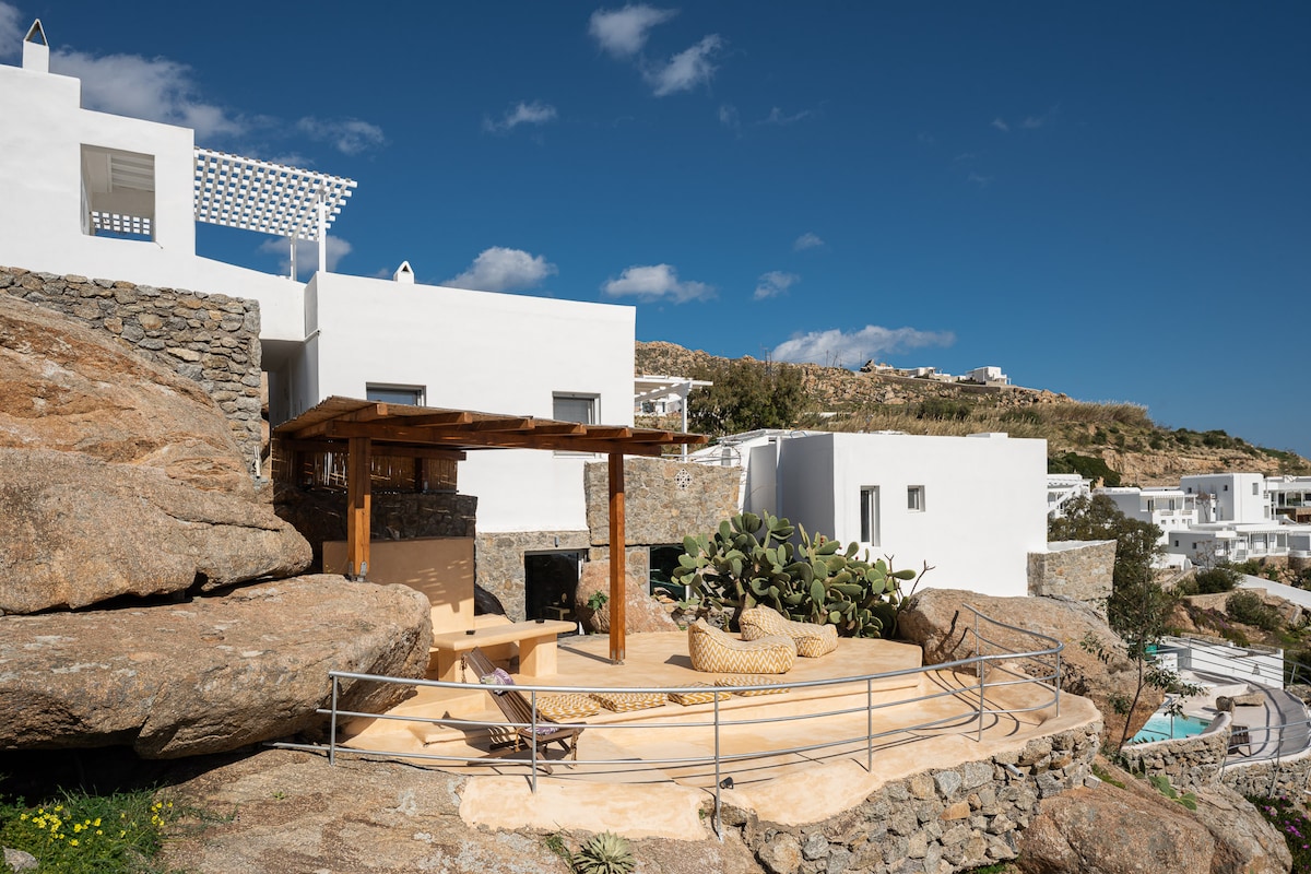 An outdoor seating area is set against natural rock formations, with a wooden pergola providing shade. Comfortable seating is positioned around a small table, surrounded by greenery. The villa's architecture is showcased with white walls in the background, harmonizing with the rocky landscape.