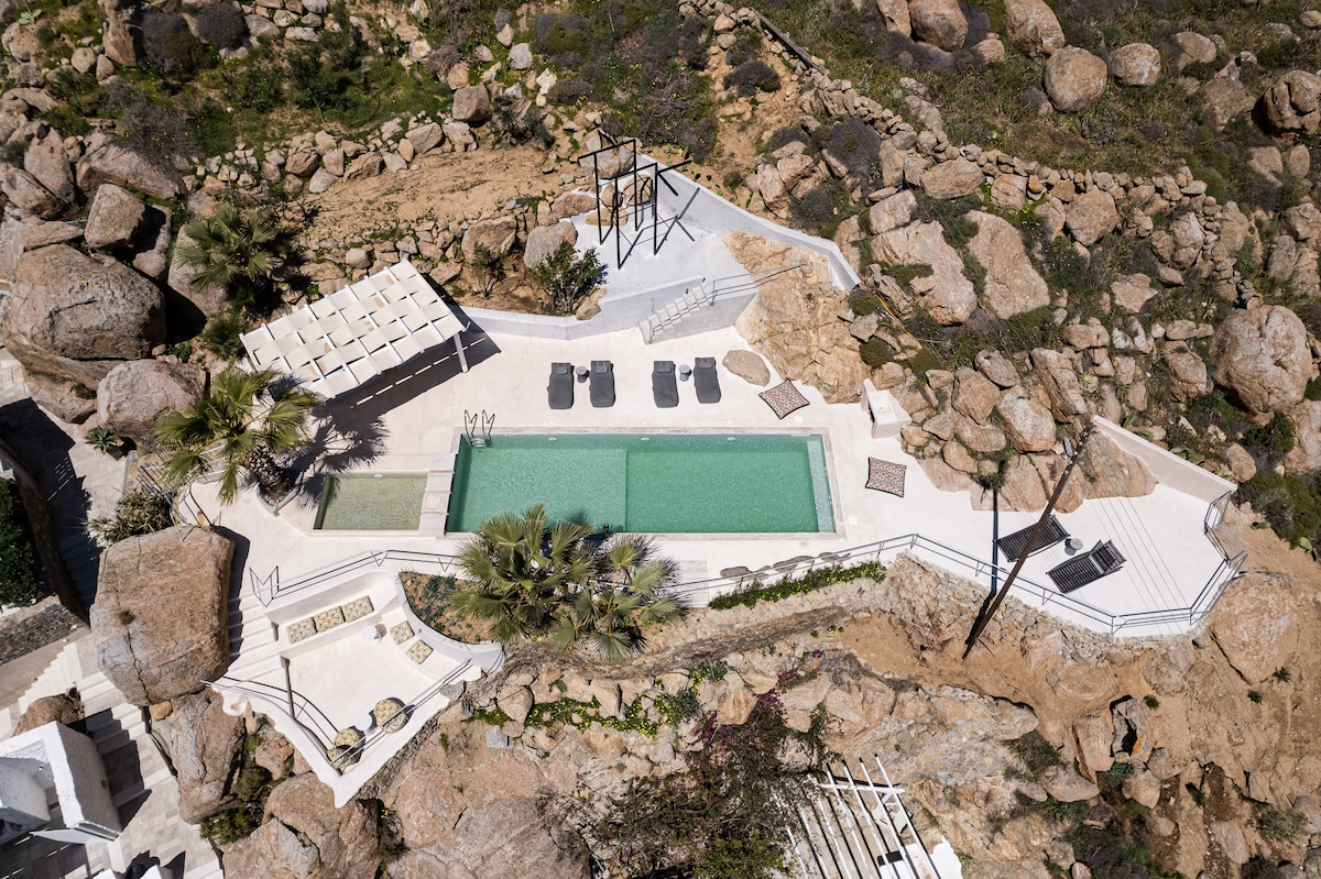 An aerial view showcases a pool area surrounded by natural rock formations. Sun loungers are arranged beside the pool, with shaded seating areas also visible. Lush vegetation contrasts with the stark rocky terrain, creating a serene outdoor environment.