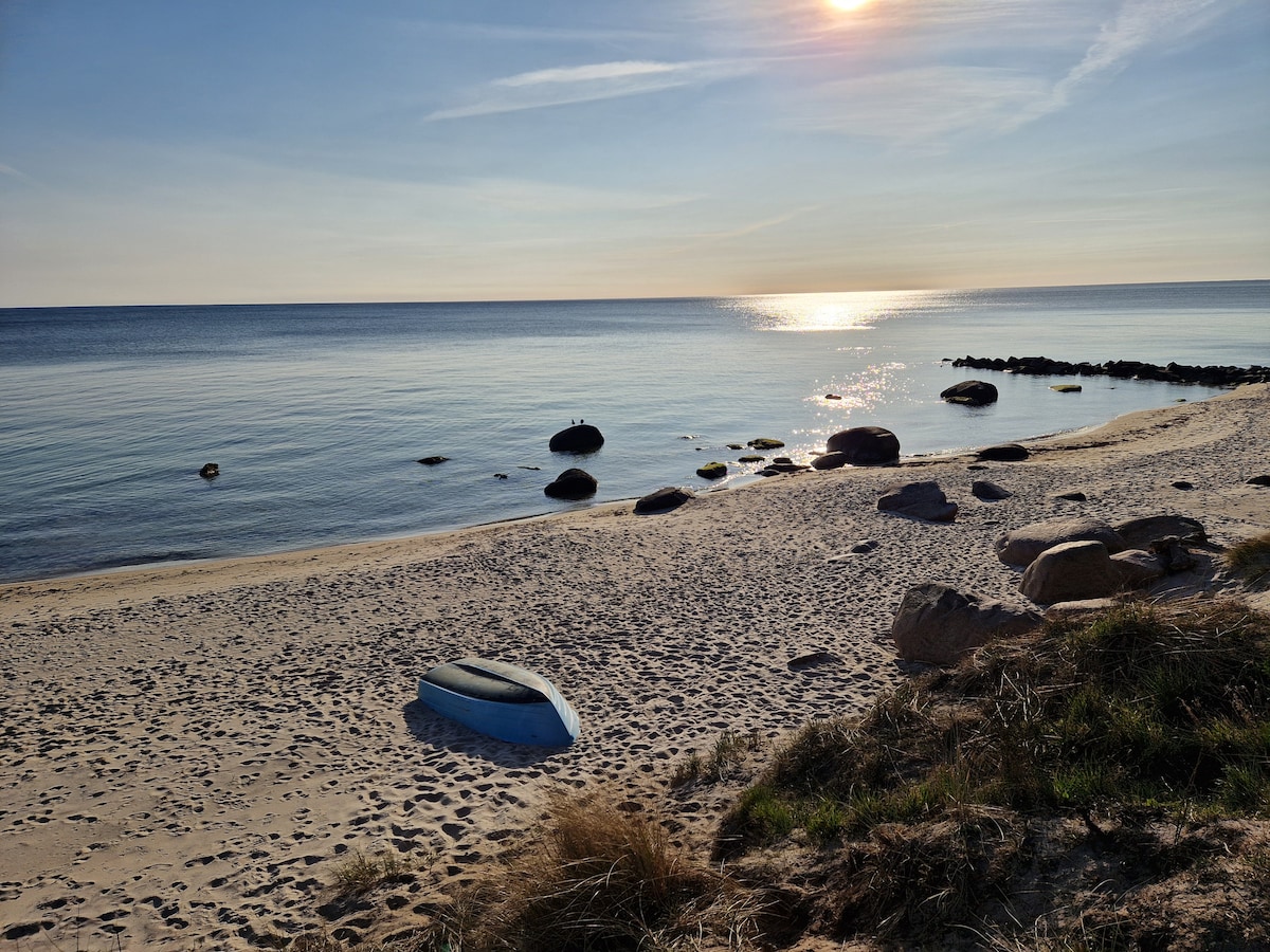 A sandy beach is viewed from a gentle rise, where a blue boat rests on the shore. Smooth stones are scattered along the waterline, and calm waves reflect the sunlight. The horizon features a clear sky, enhancing the serene coastal ambiance.