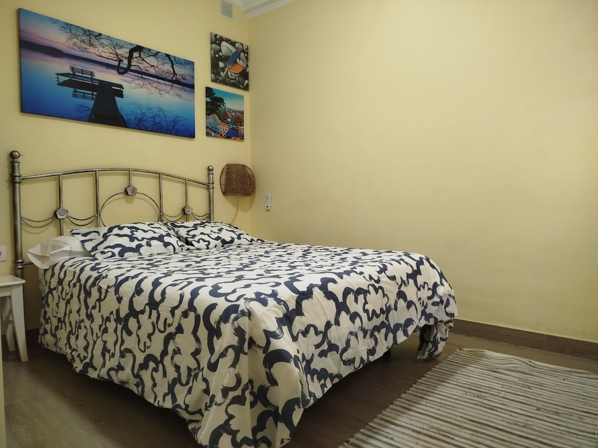 A spacious bedroom features a large bed with a patterned bedspread in blue and white. Two artworks are hung on the yellow wall above, showcasing scenic landscapes. Light streams in, creating a warm ambiance, while a woven wall hanging adds texture to the room.