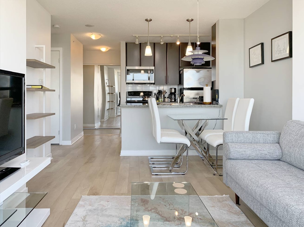 An open-concept living space is showcased, featuring a modern kitchen with stainless steel appliances and granite countertops. A sleek dining area is visible with a glass table and white chairs, while a grey sofa complements the light-colored flooring and neutral walls.