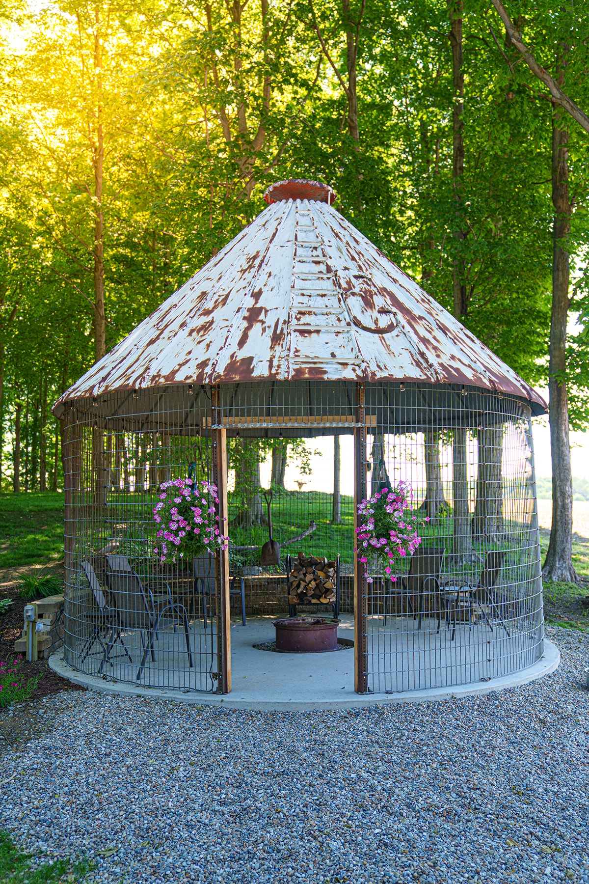 A unique gazebo with a rusted metal roof is surrounded by lush trees, providing a sheltered space for relaxation. Four chairs are arranged around a fire pit in the center, and flower baskets hang from the sides, adding color to the peaceful outdoor setting.