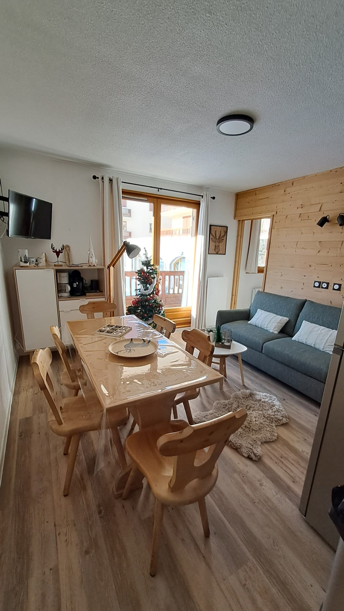 A dining area features a long wooden table surrounded by six light-colored chairs. A small decorative tree is displayed on the table. A comfortable gray sofa and a wall-mounted television are visible in the living space, with large windows allowing natural light to fill the room.