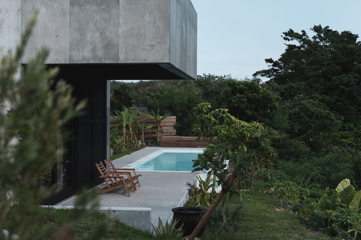 An outdoor area features a rectangular infinity pool surrounded by greenery. Two wooden deck chairs are positioned near the pool, offering a tranquil space for relaxation. The structure above has a modern concrete facade, blending with the natural landscape.
