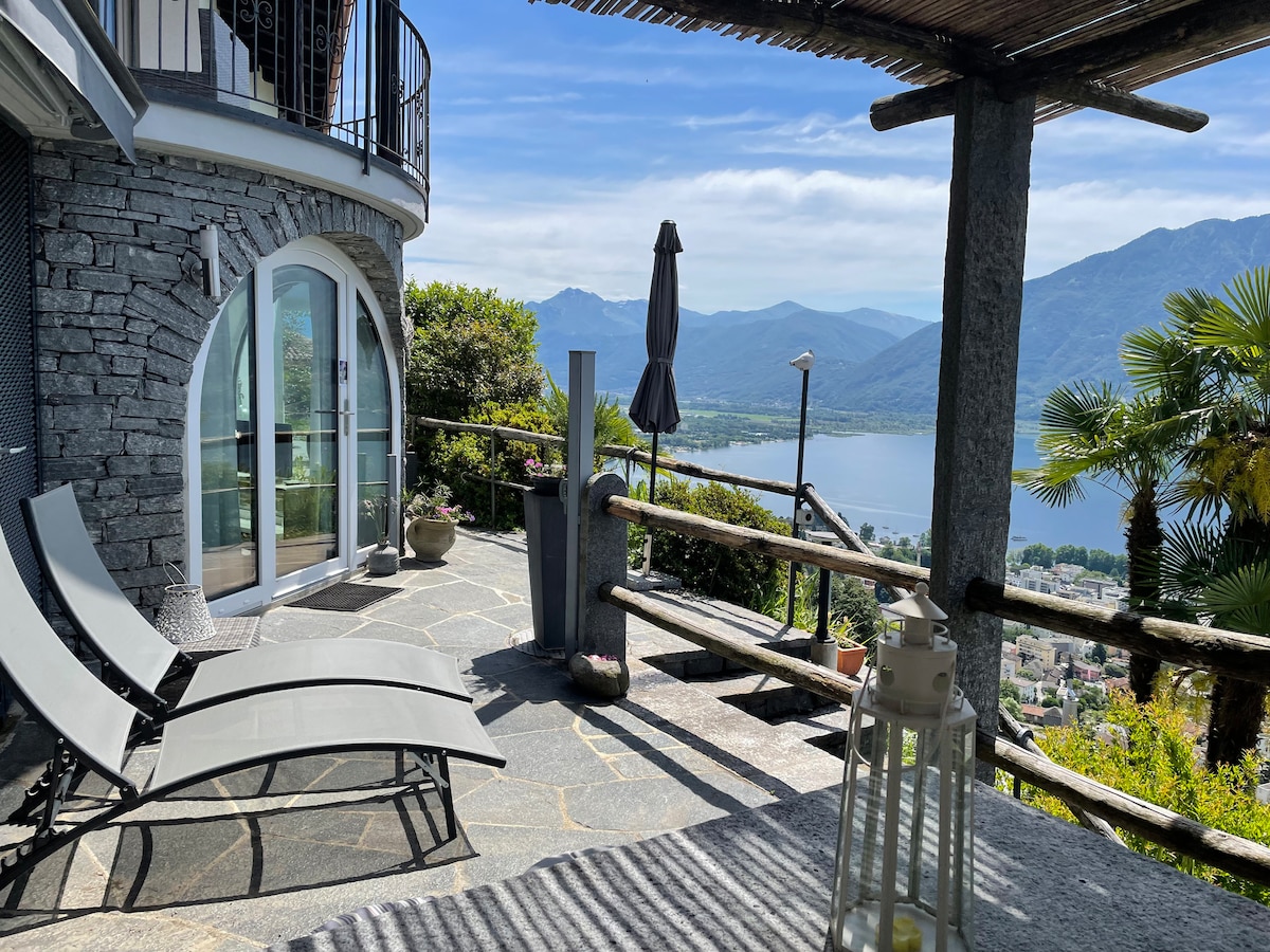 An outdoor terrace is displayed with two gray lounge chairs positioned on natural stone flooring. A shaded umbrella stands nearby, and a lantern adds a touch of ambiance. Scenic views of the lake and mountains are visible in the background.