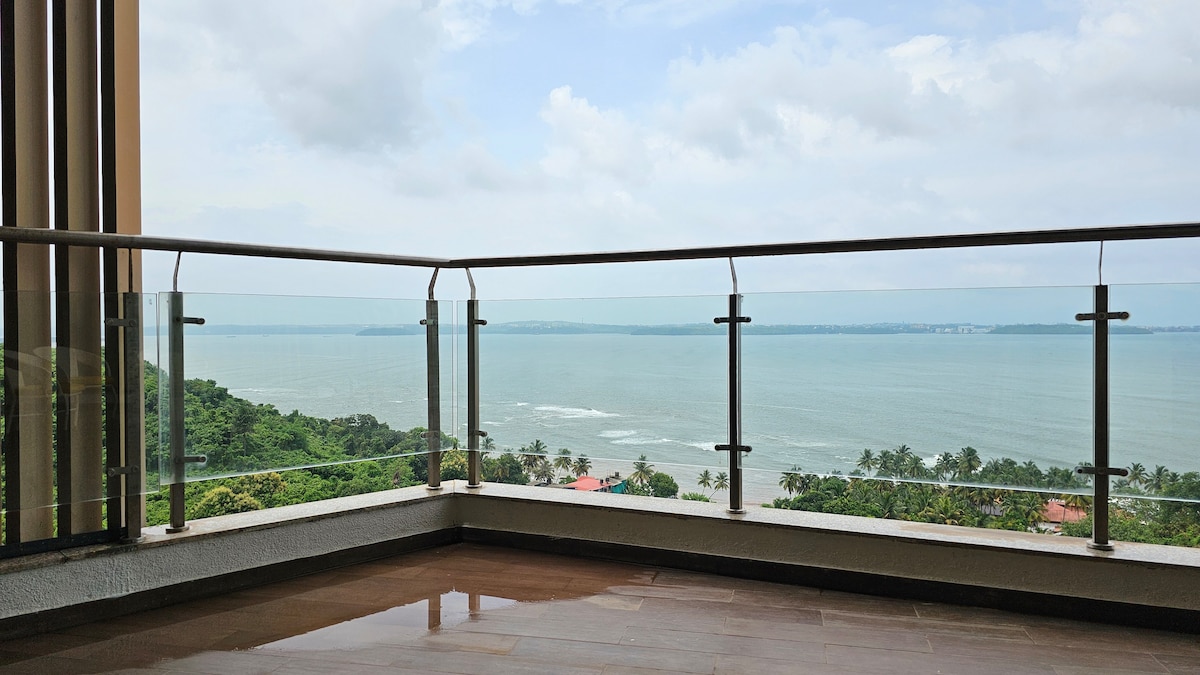 A spacious balcony offers stunning sea views, framed by glass railings. The tranquil blue waters reflect the sky, while the surrounding greenery enhances the picturesque setting. The floor is tiled, providing a clean and inviting space for relaxation.