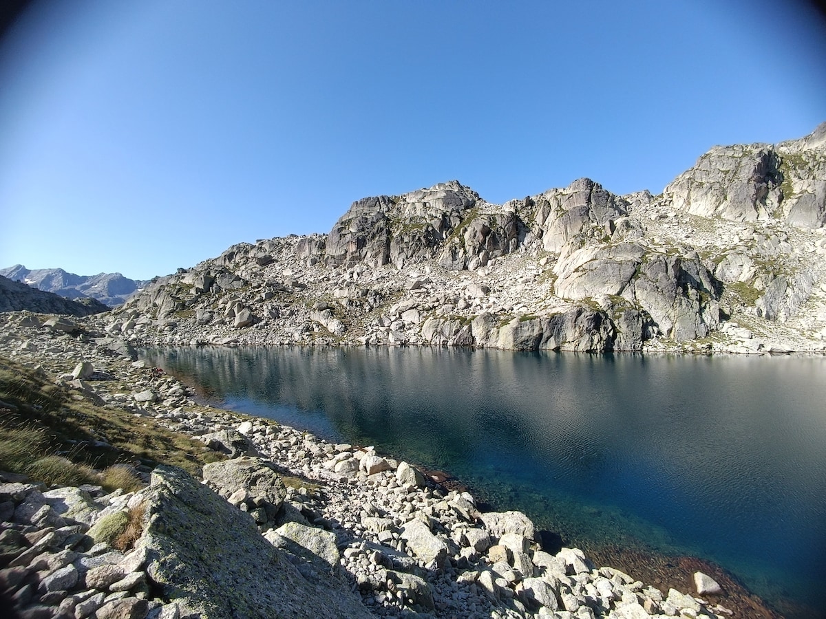 A serene alpine lake is framed by rugged rocky terrain under a clear blue sky. The still water reflects the surrounding mountains, while patches of greenery and scattered boulders offer a natural contrast. The tranquil environment is ideal for enjoying nature.