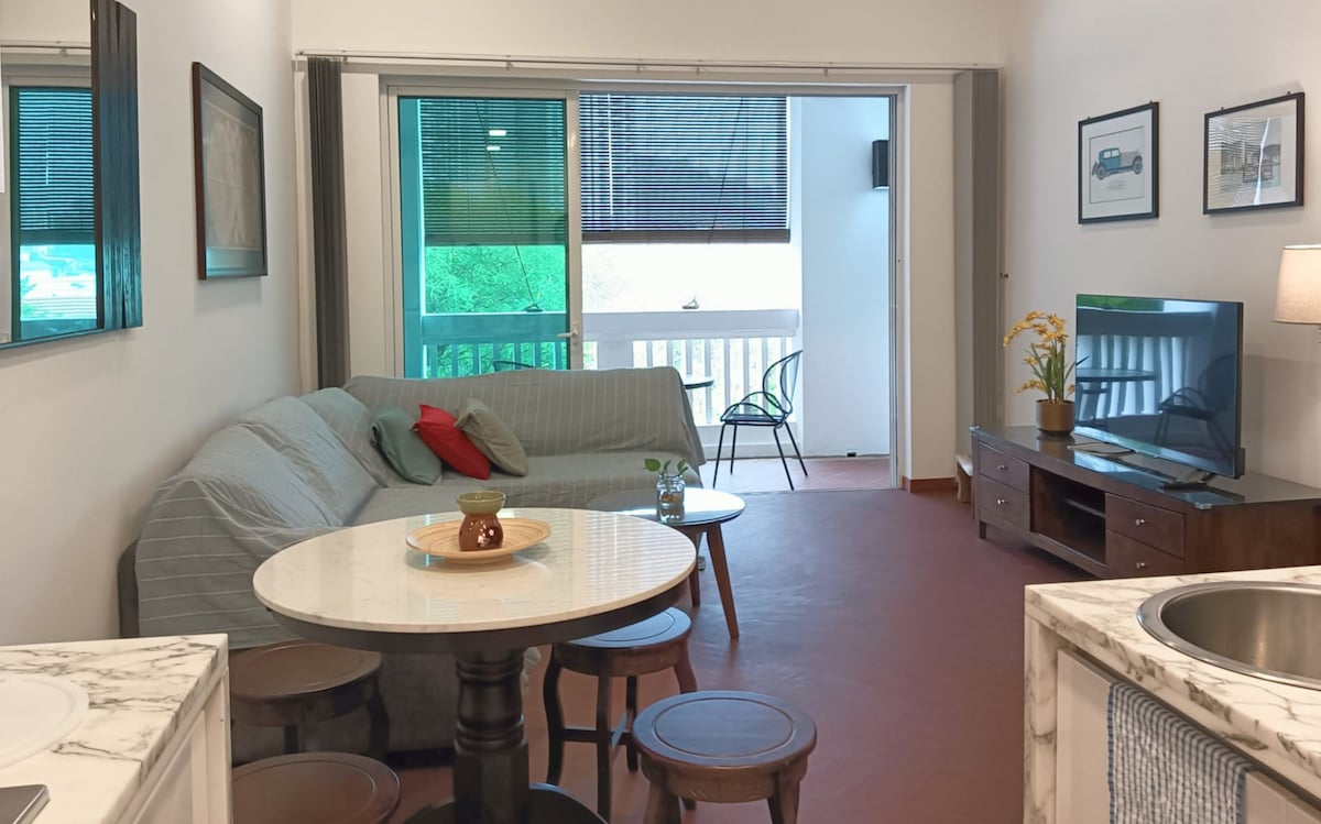 A living area features a comfortable sofa with colorful cushions and a round dining table with stools. Natural light filters through large sliding glass doors that open to a private balcony, providing a view of greenery. A television is positioned on a wooden media console.