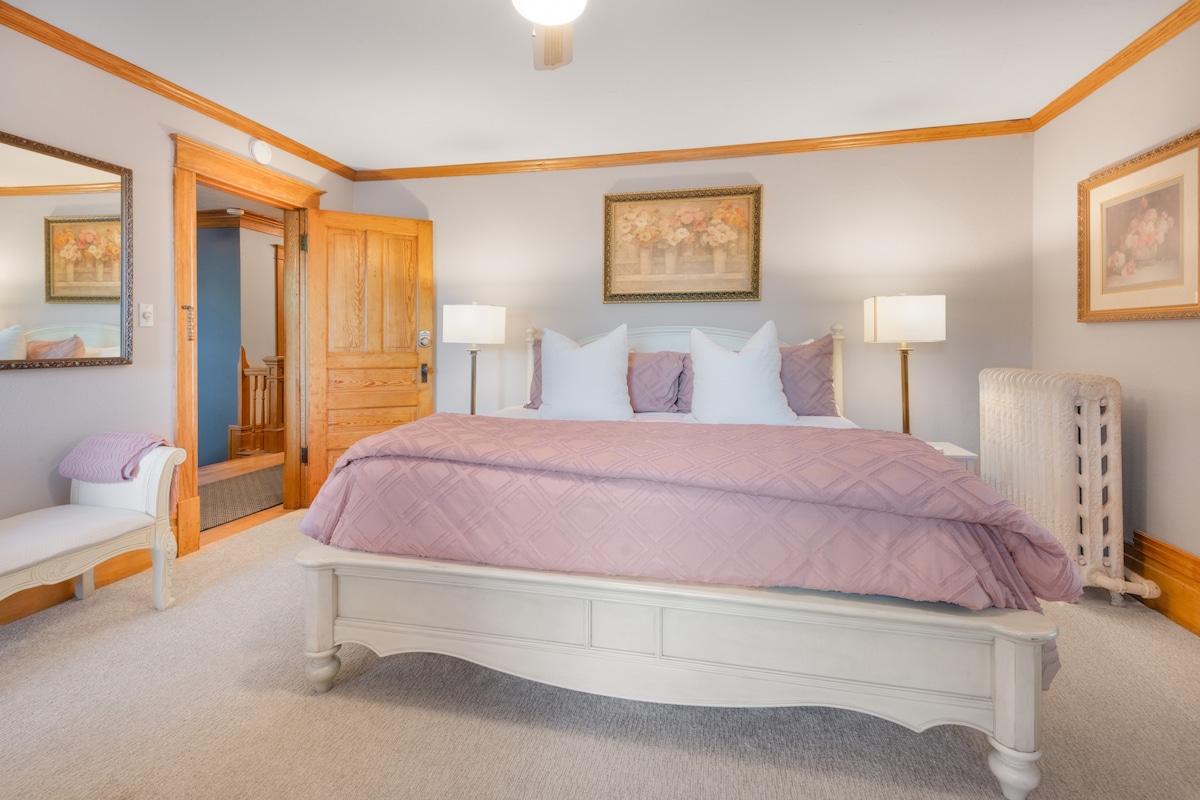 A spacious room showcases a King-sized bed dressed in soft pink bedding. Two bedside lamps flank the bed, providing gentle illumination. Light-colored trim and decorative artwork accent the walls, while a vintage radiator contributes to the room's historical charm. A door leads to the hallway.