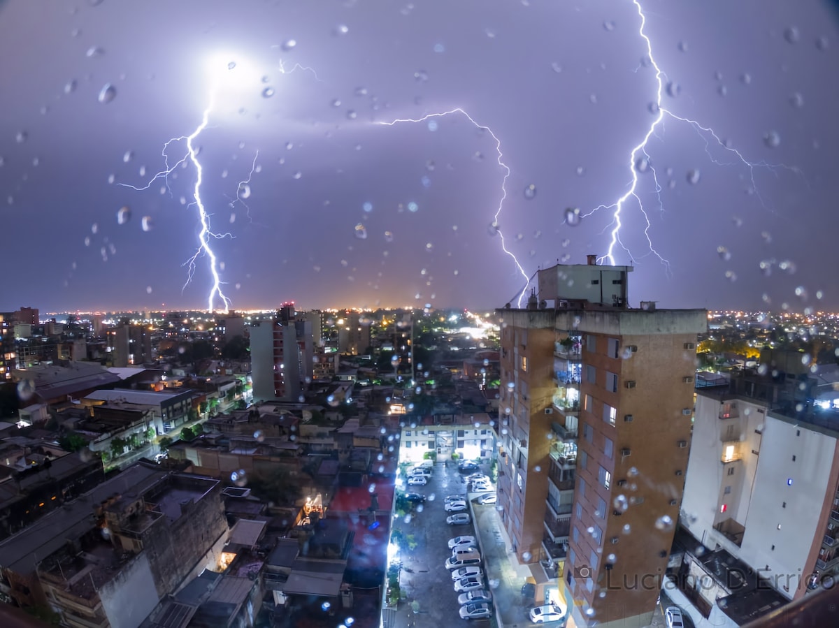 A dramatic cityscape is captured during a thunderstorm, with bright lightning streaks illuminating the sky. Raindrops are visible on the glass, creating a blurred effect, while city lights shine below, contrasting with the cloudy atmosphere.