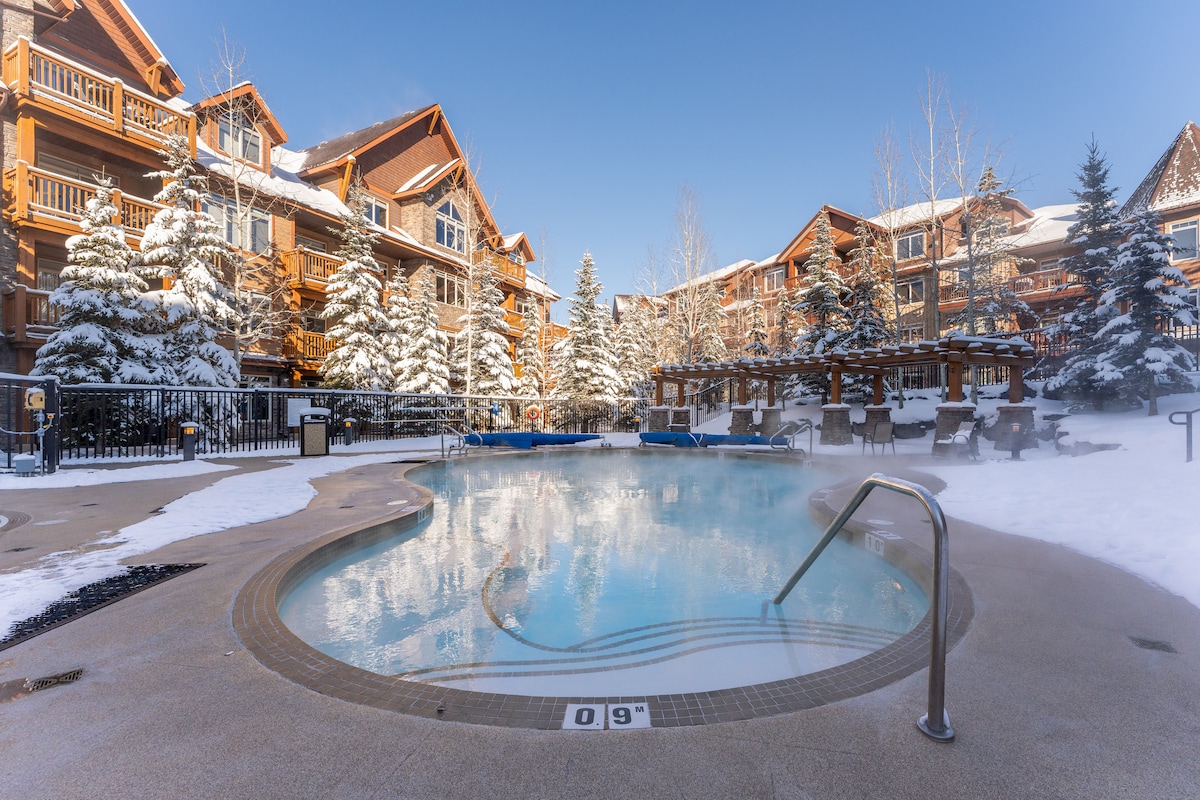 An outdoor heated pool is surrounded by snow-covered trees and charming wooden buildings. Steam rises gently from the water, creating a serene atmosphere. Loungers are arranged invitingly around the pool, offering a cozy spot to relax while enjoying the winter scenery.
