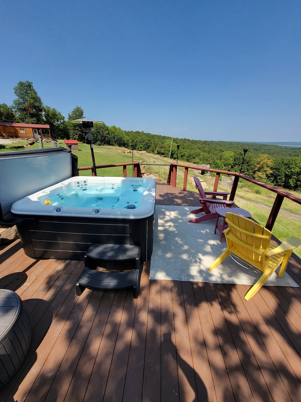 A hot tub is situated on a wooden deck, providing views of the surrounding landscape. Colorful chairs in red and yellow are placed nearby, offering spaces to relax. Sunlight casts soft shadows on the deck, enhancing the peaceful outdoor setting.