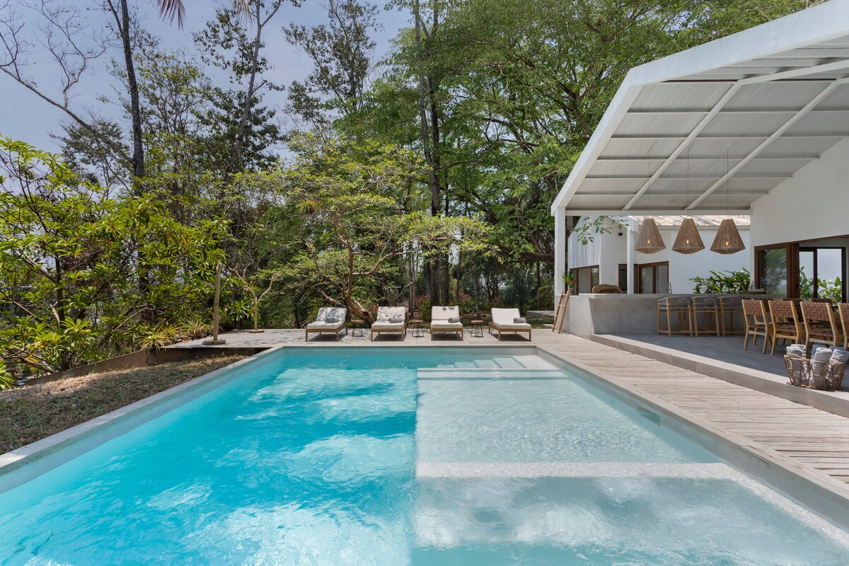 A tranquil outdoor area features a sparkling pool surrounded by wooden decking. Several lounge chairs are positioned for relaxation under the shade of trees. A covered seating area with pendant lighting and a bar counter is visible in the background, extending the space into nature.
