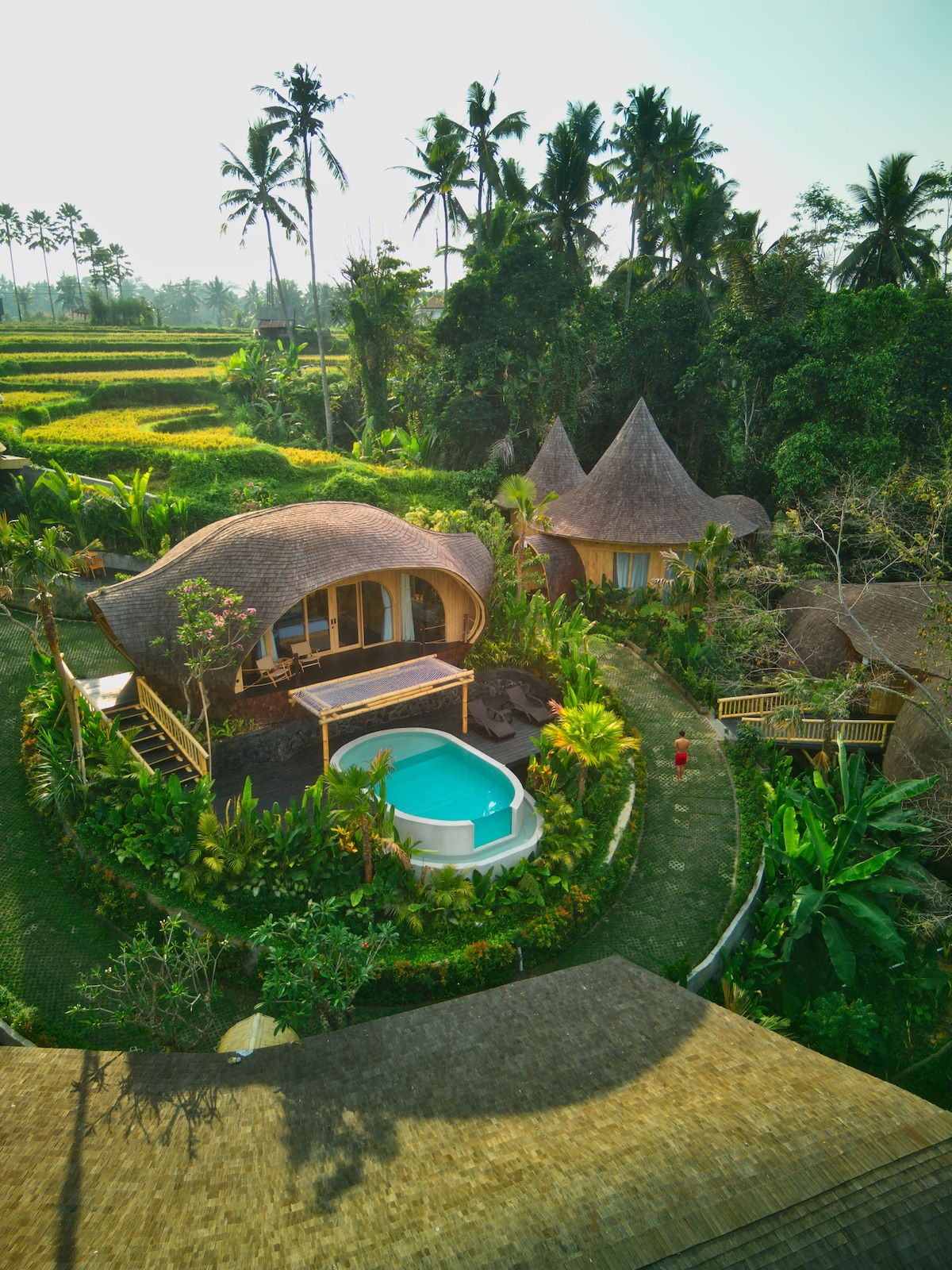 An aerial view of a unique bamboo house surrounded by lush greenery and tropical plants. A private pool is visible in the outdoor area, complemented by a cozy patio. The distinct, pointed roofs of the nearby structures add charm to the serene landscape.