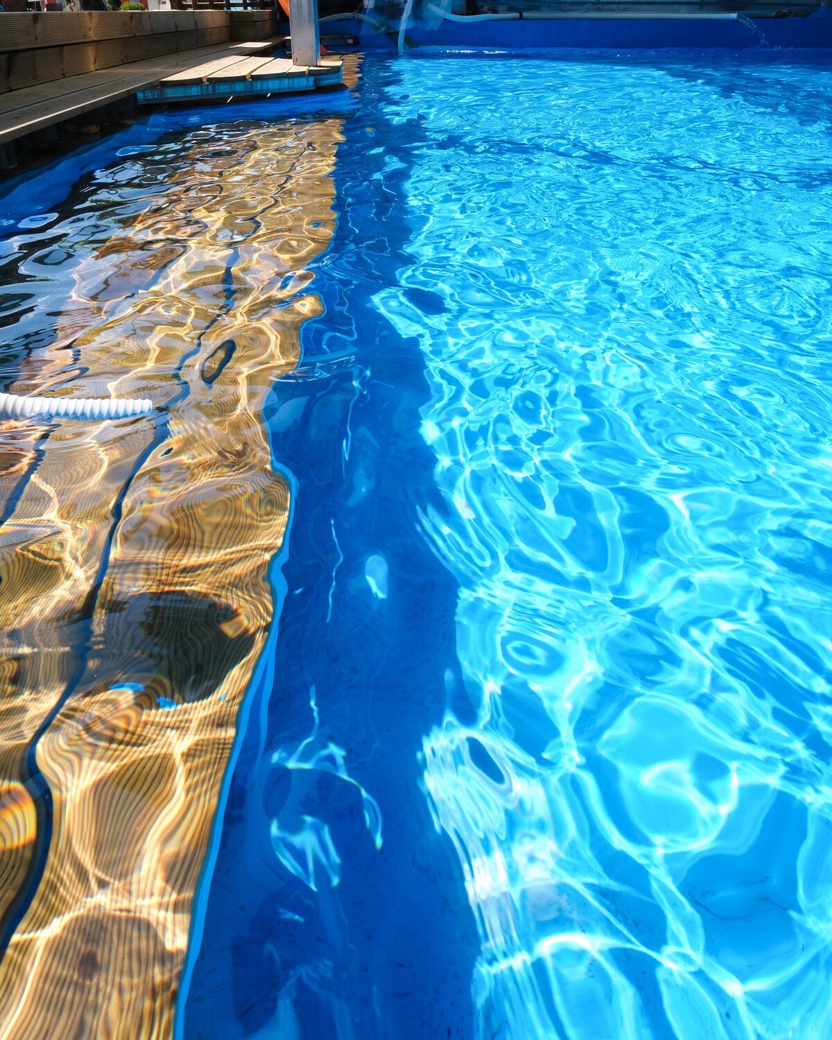 A clear swimming pool is visible, with sunlight creating a shimmering effect on the water's surface. The reflection of wooden decking can be seen along the edge, complementing the vibrant blue hue of the pool.