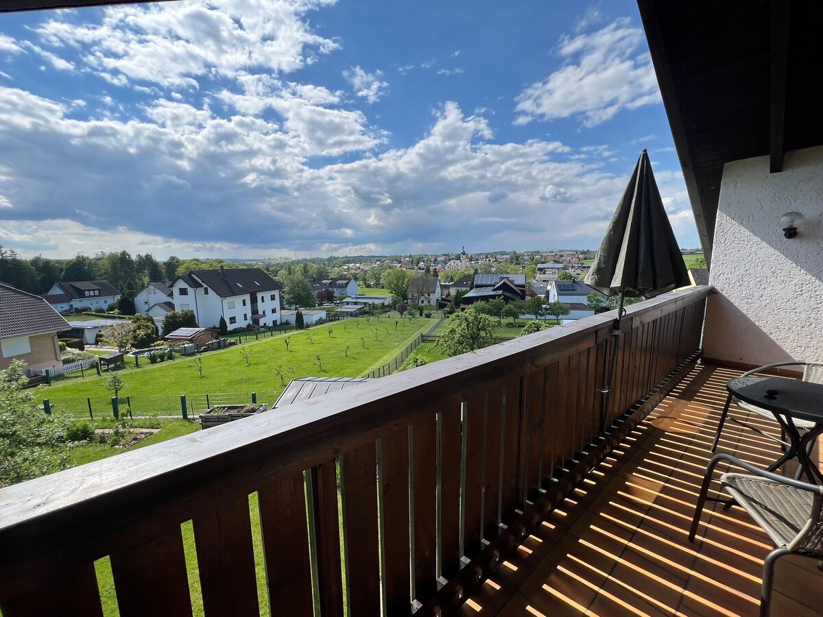 A balcony with wooden railing offers a scenic view of the surrounding countryside, including green fields and distant houses. The space features a small table and chairs, with a shaded area provided by a striped umbrella. Soft clouds drift across the blue sky.