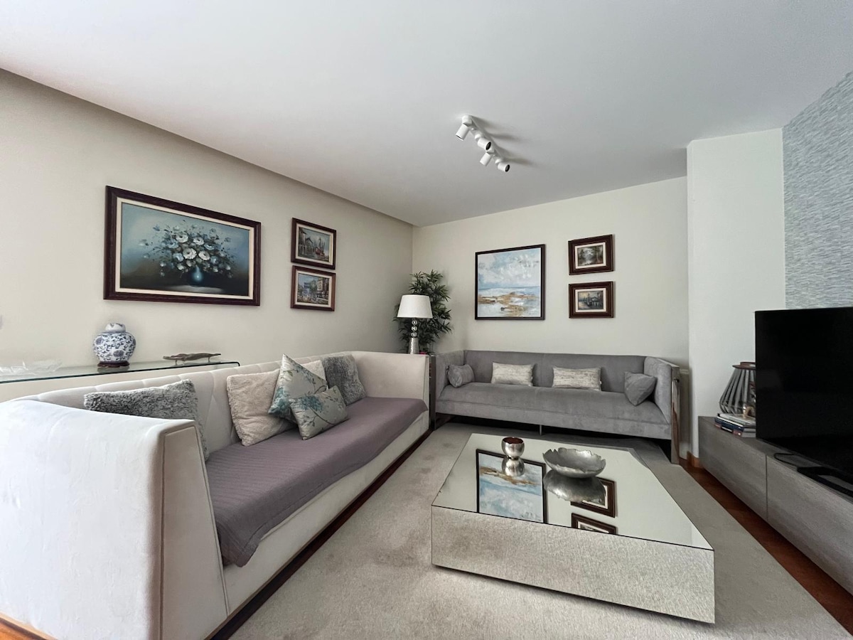 A sophisticated living room features a light-colored sectional sofa paired with a matching gray couch. An elegant glass coffee table reflects the room's soft lighting. Wall art is displayed in framed arrangements, and a decorative plant adds a touch of greenery.
